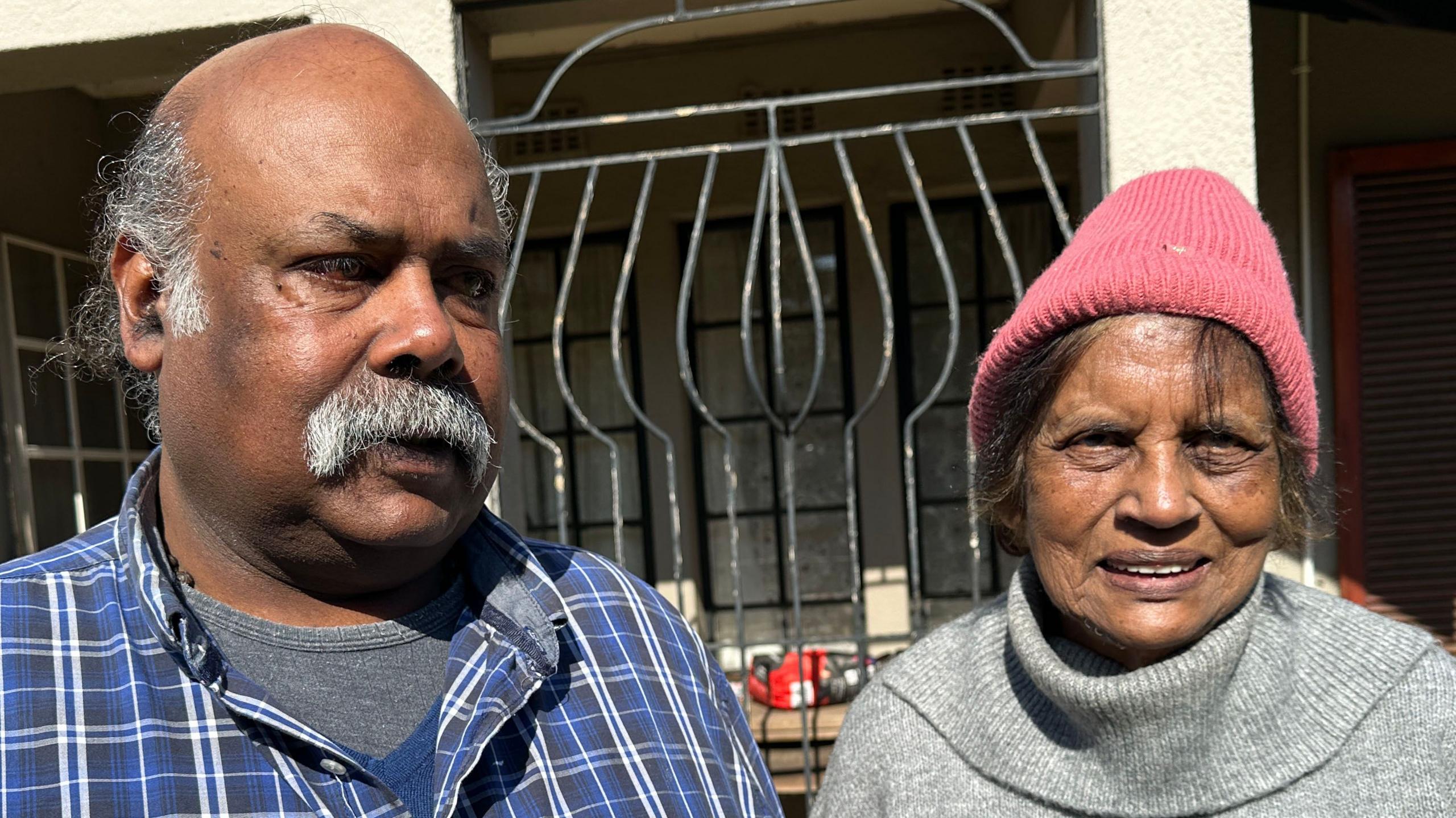 Mark Moodley stands next to his mother. He is wearing a blue-checked shirt and she is wearing a grey jumper and pink beanie hat.