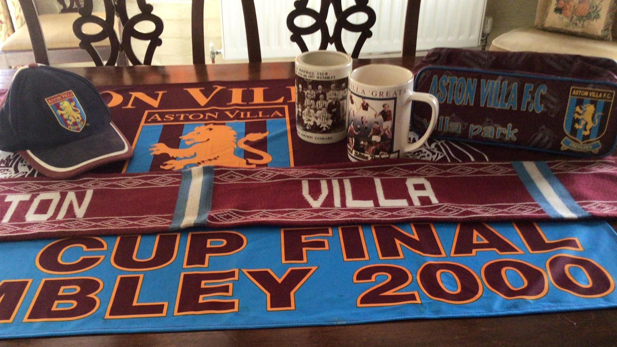 Aston Villa scarfs, cups and caps are sitting on a table. They are red, blue and orange in colour