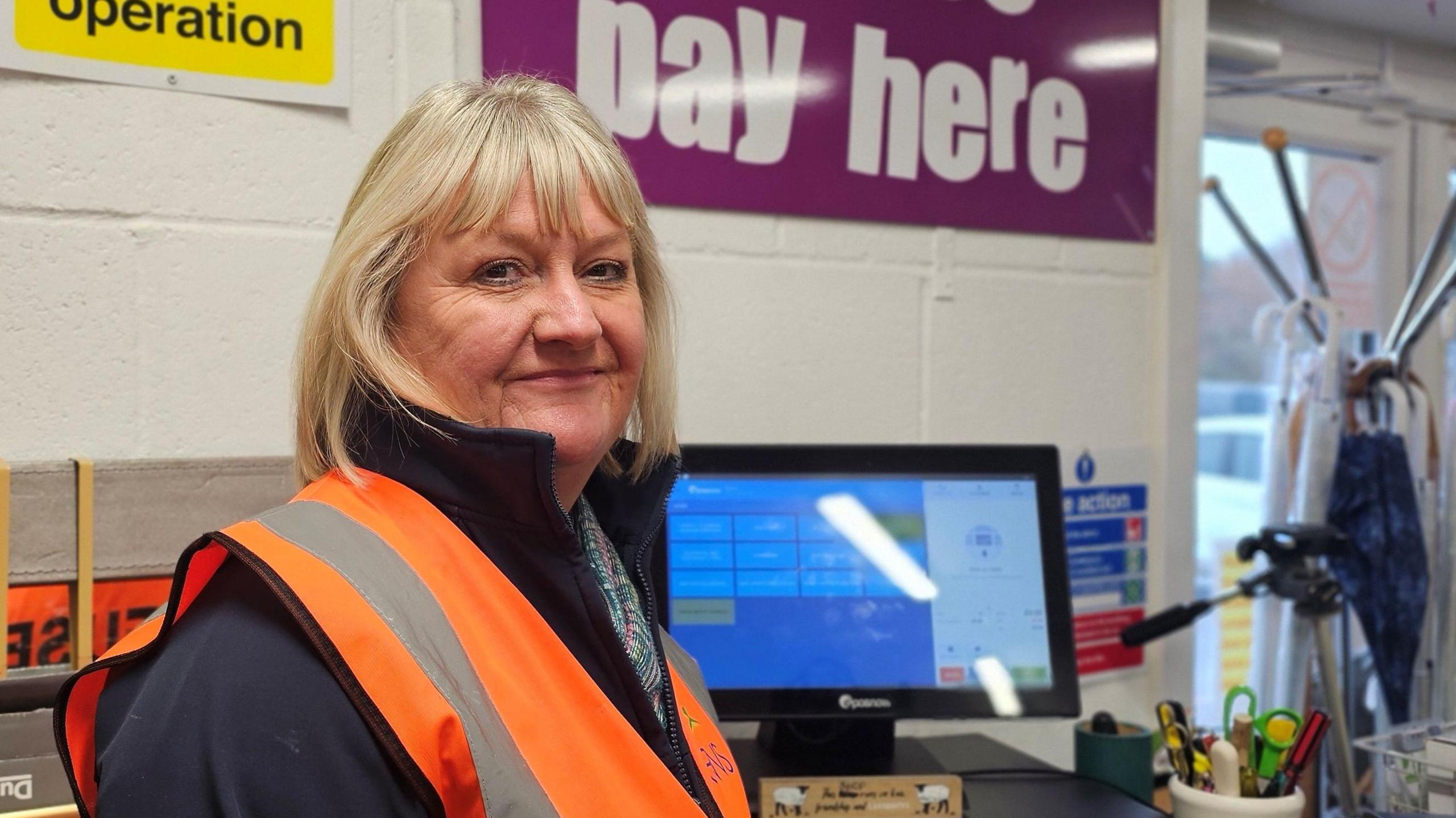 Karen Betts is smiling at the camera, She's wearing an orange high-vis waist over a dark blue fleece. Behind her, we can see the till and a sign saying "pay here"