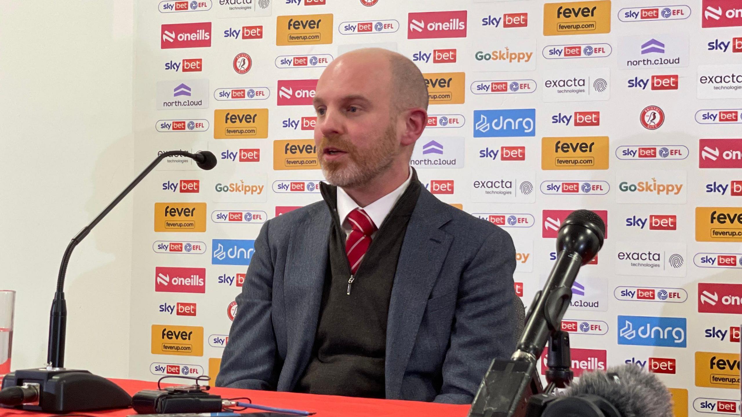 Charlie Boss sitting in front of a table with microphones at a press conference. He is wearing a blue suit jacket, red tie, white shirt and black jumper