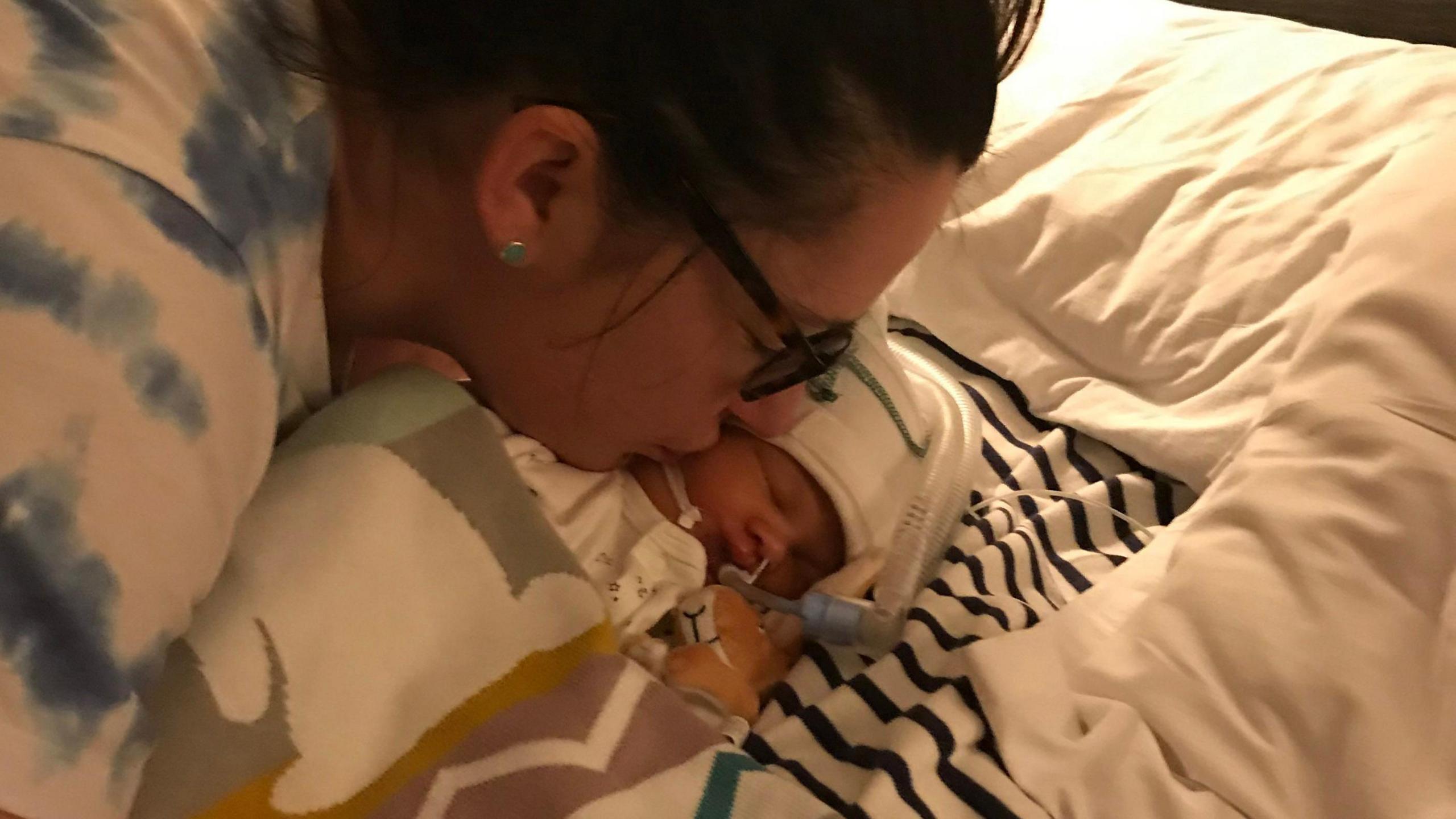 Sarah Robinson, who is wearing glasses and has dark hair tied back in a ponytail, kiss the cheek of newborn Ida Lock, in a hospital bed.