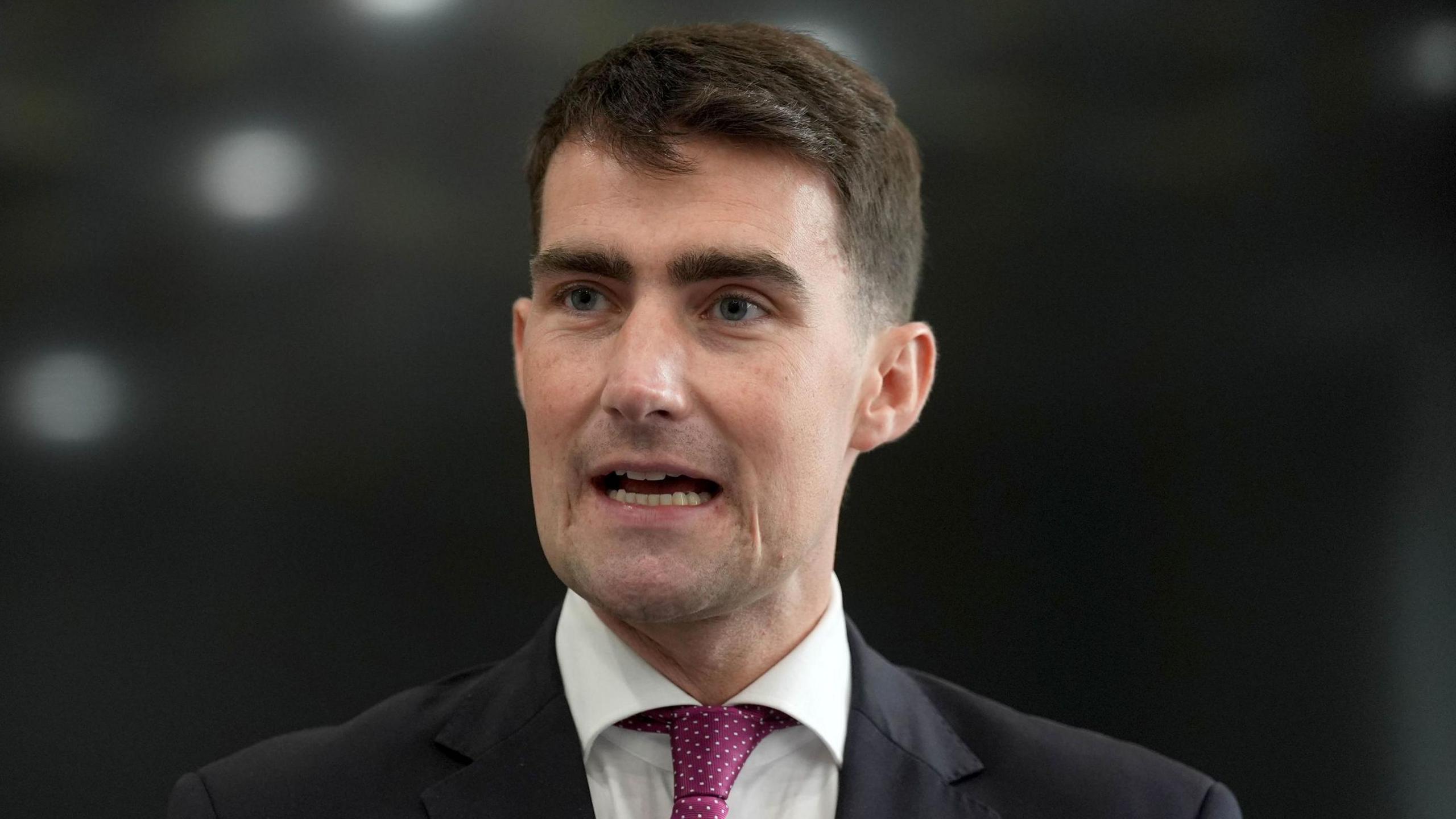 Jack Chambers is wearing a suit, white shirt and pink tie. He has short black hair. The background is blurry. 