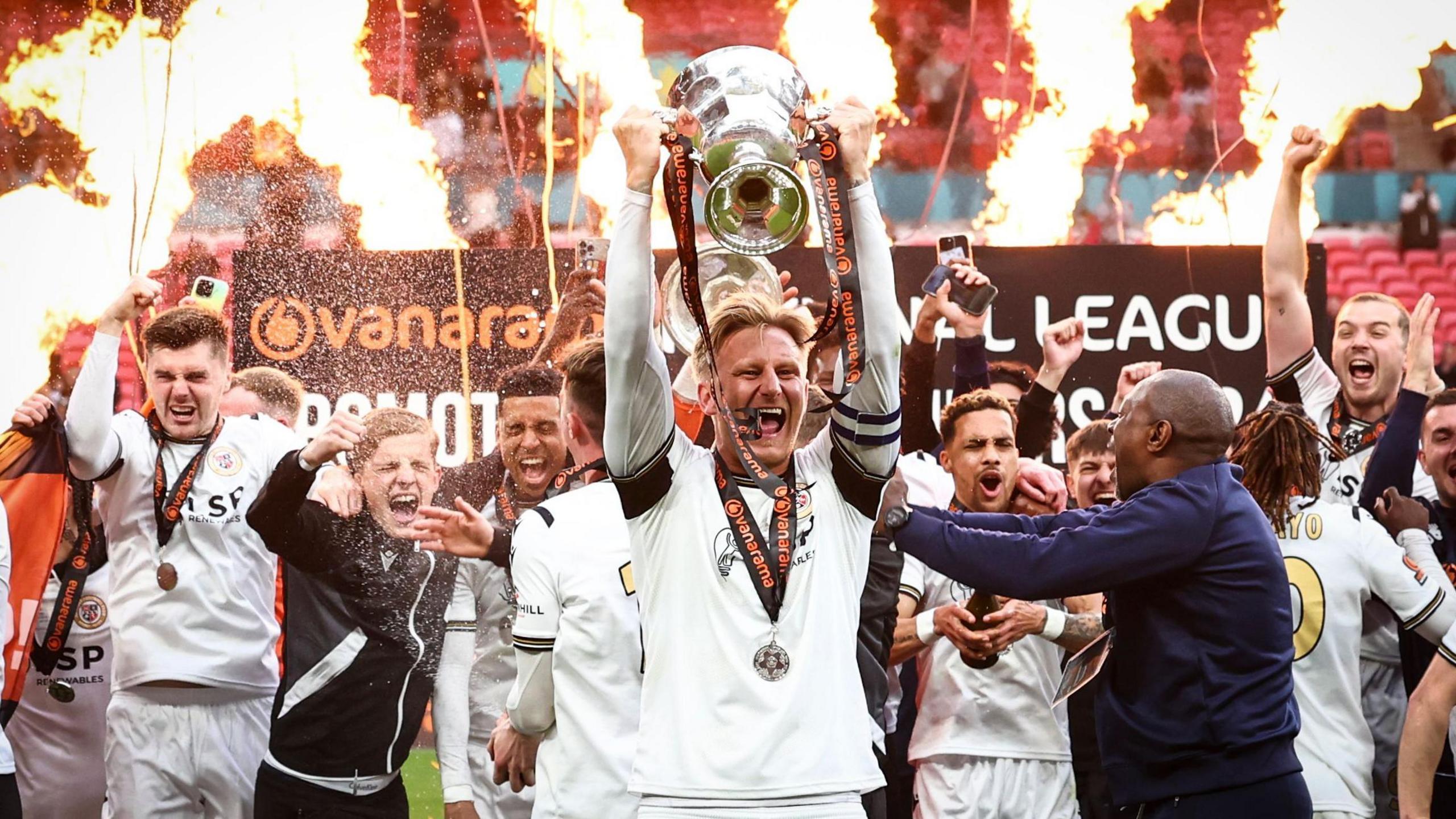 Captain Byron Webster holds the National League promotion final trophy aloft in front of celebrating Bromley players