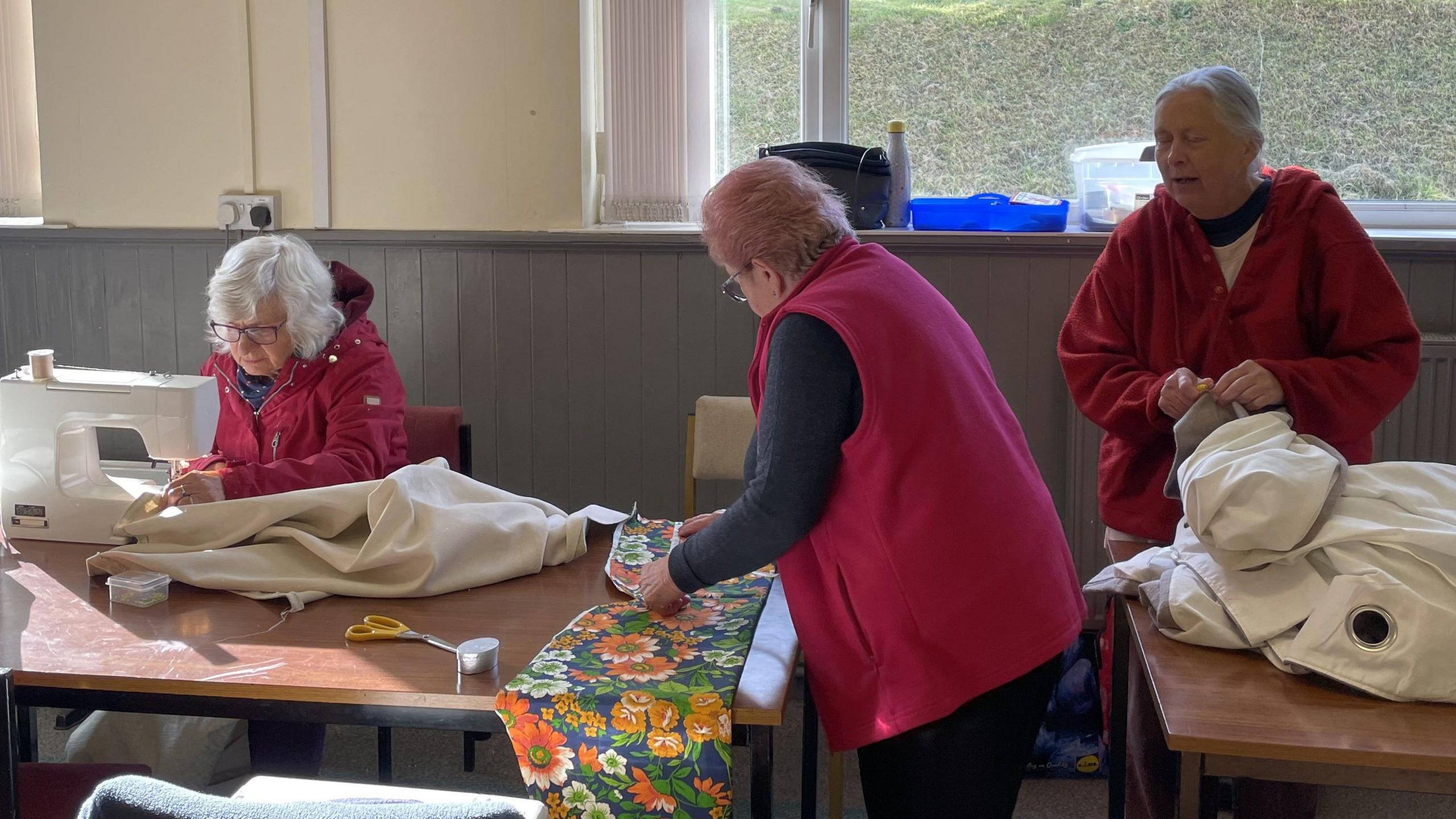 A number of women using sewing machines prepare materials on long wooden tables