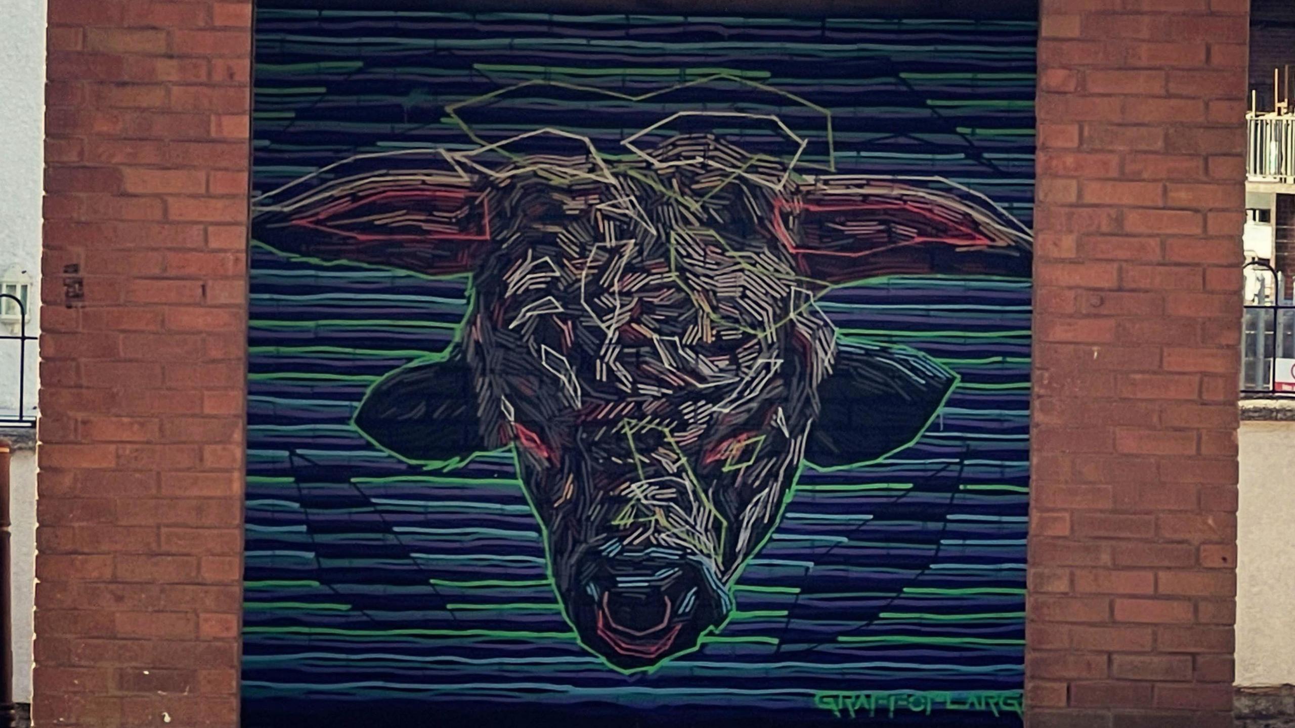 A bull head painted in lines which include green, pink and white. There are also horizontal lines against a blue background and the design is against a brick wall. 