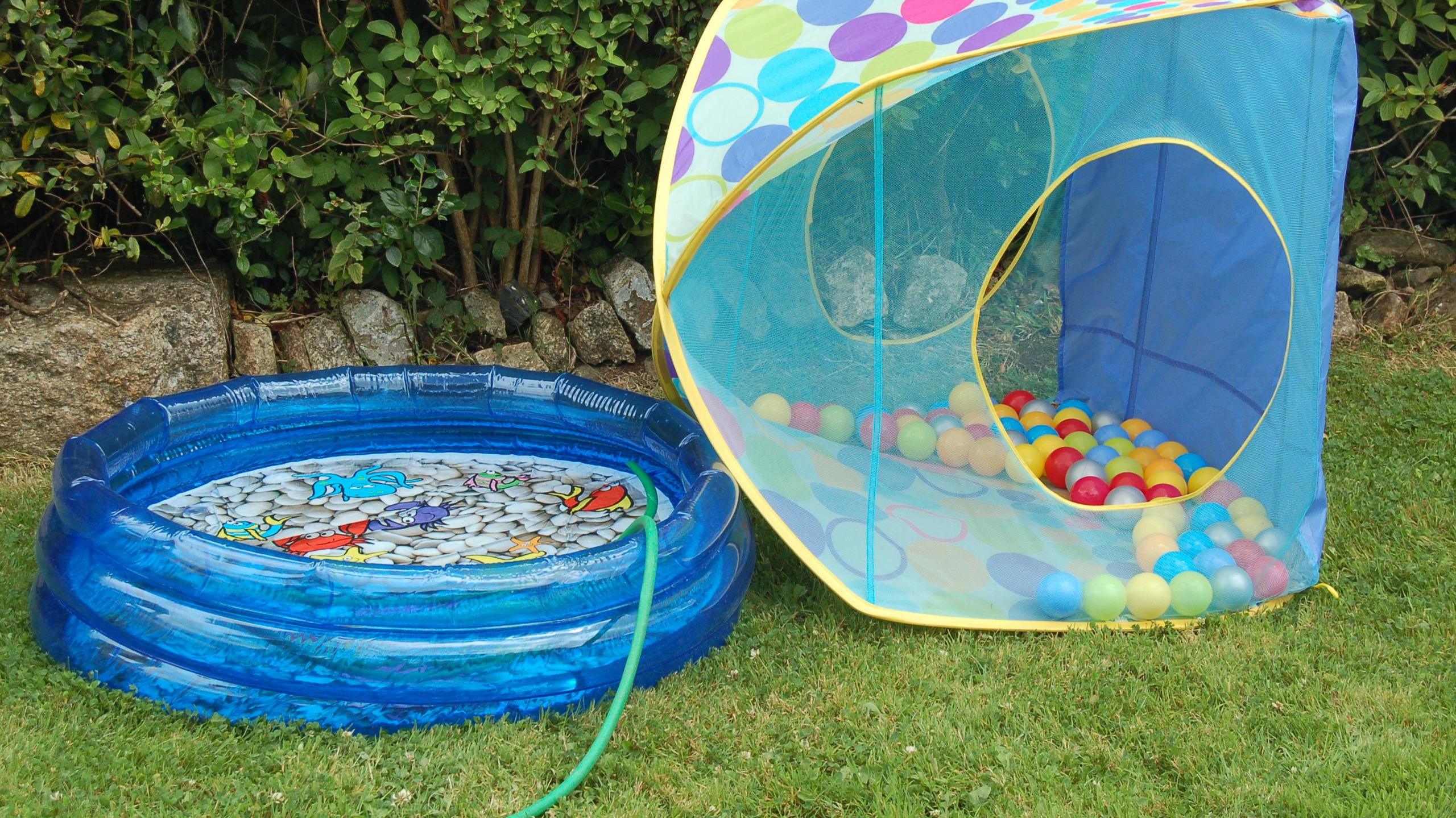 A blue plastic paddling pool in the garden, with a green hose running into it. It is  next to a mini tent full of plastic lightweight balls.