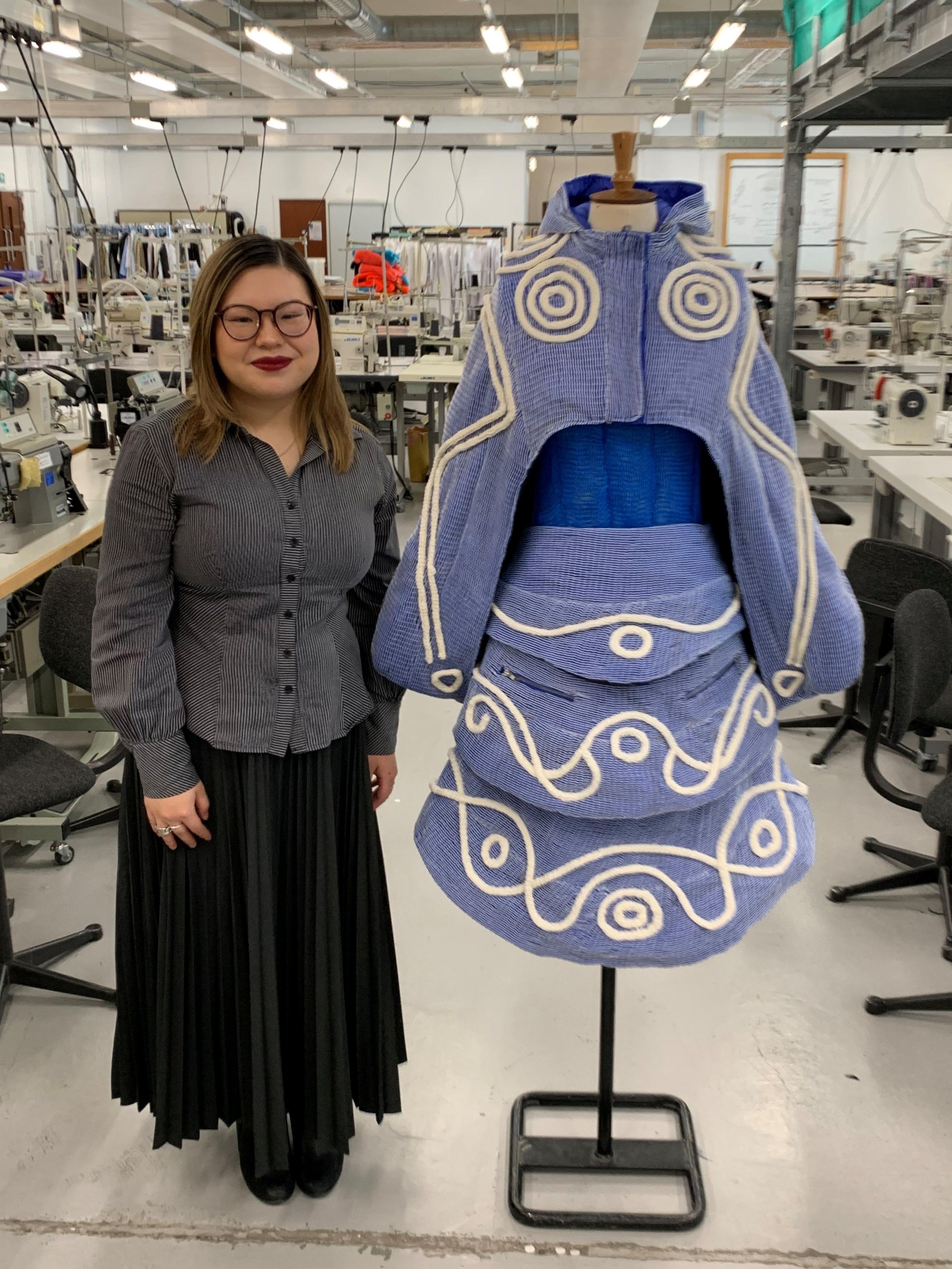 A woman in glasses with long hair in a grey blouse and black skirt stands next to a blue outfit with white embroidery
