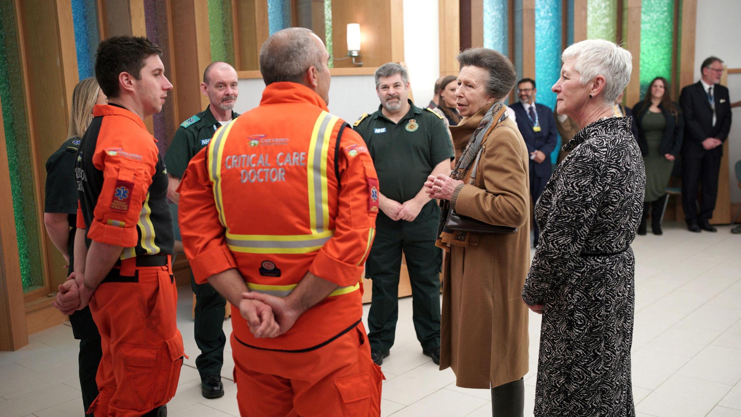 Princess Anne talks to critical care staff who are wearing orange medical scrubs in the main atrium at Southmead Hospital in Bristol. She is wearing a brown jacket and patterned scarf and other hospital staff are visible in the background