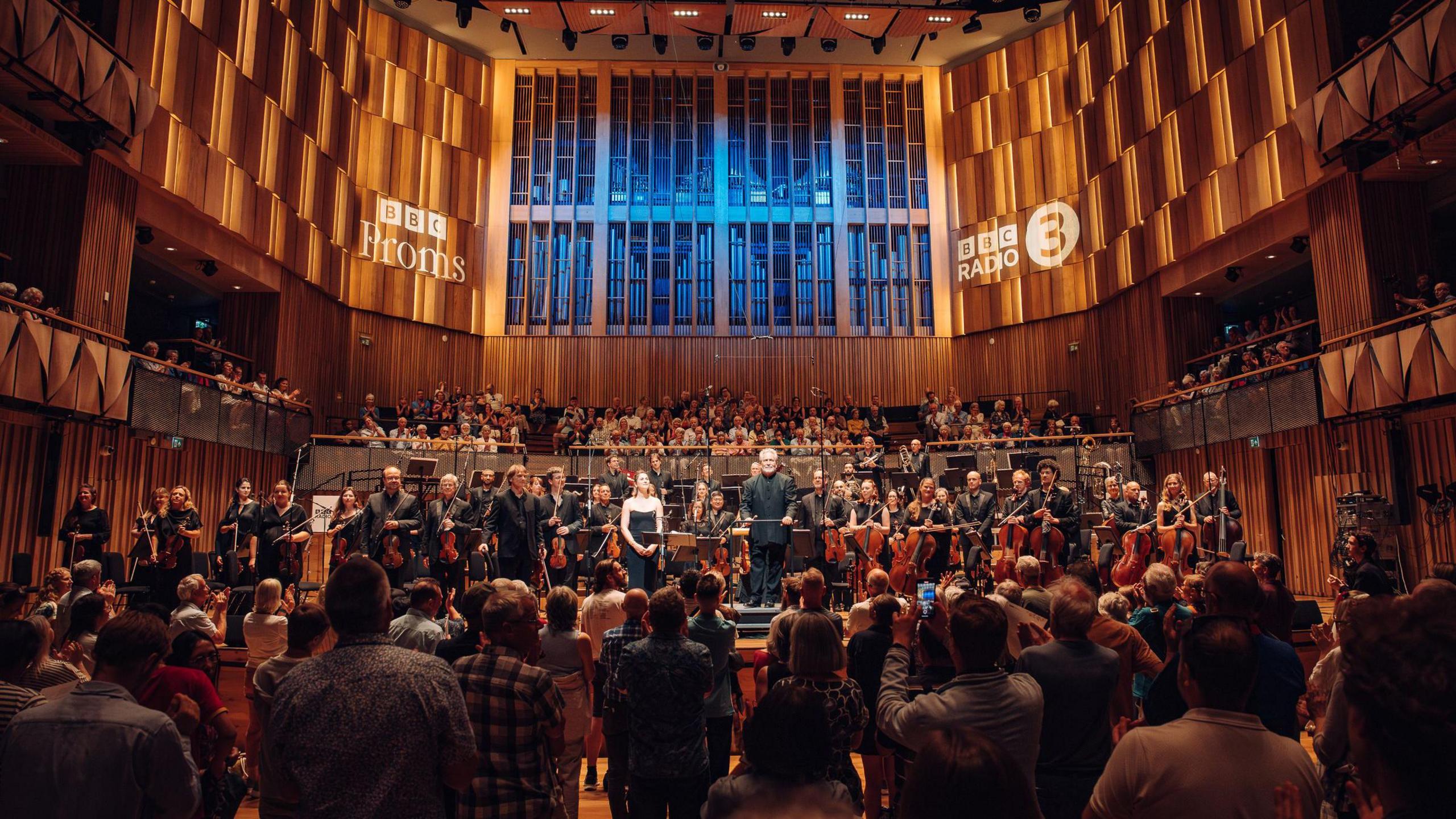 An orchestra stands on stage at the Bristol Beacon as part of the 2025 BBC Proms. On the wall behind them the text "BBC Radio 3" has been projected in two places. There is a standing audience watching the orchestra.