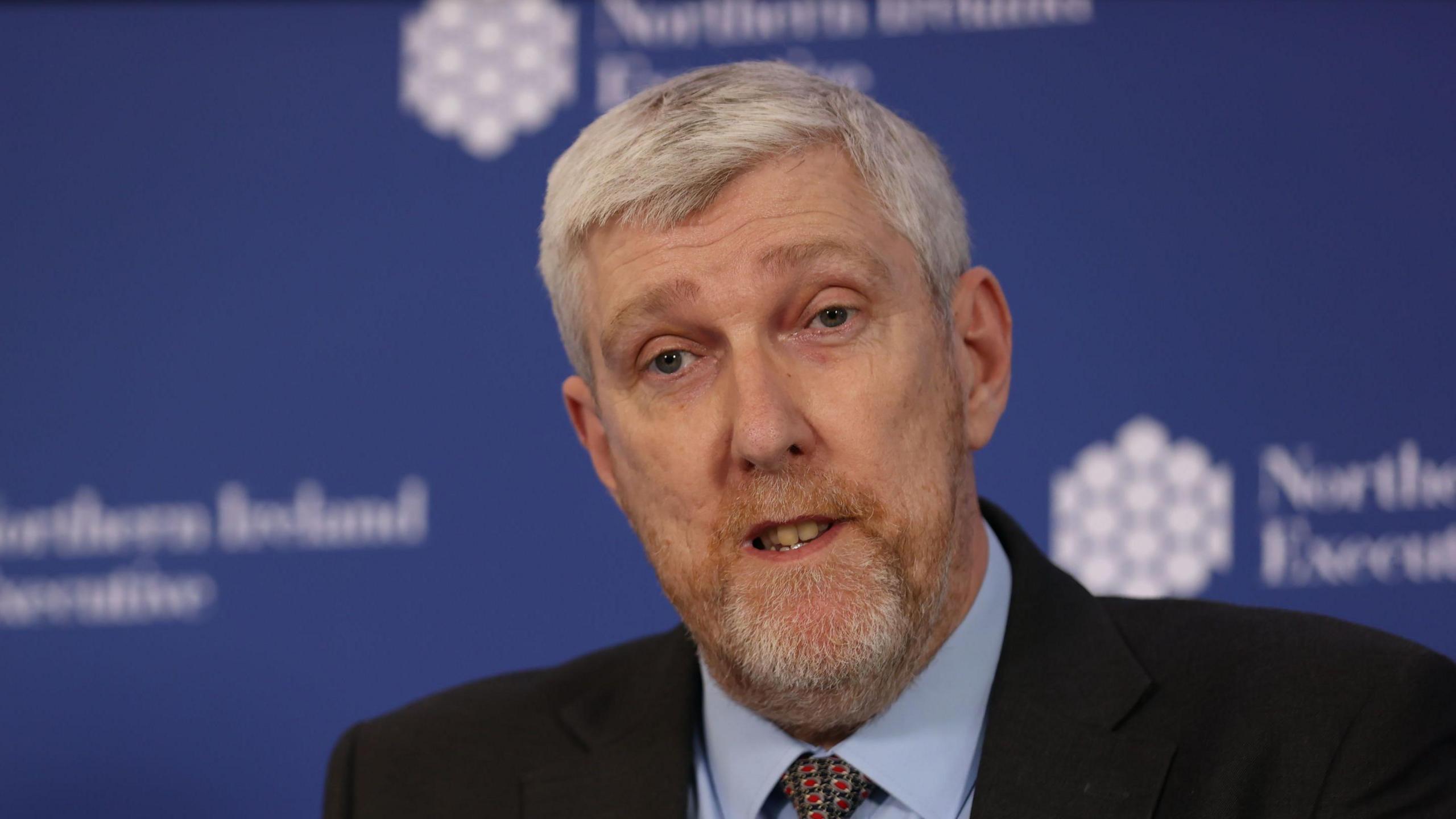 John O'Dowd is wearing a blue shirt with a red and green tie. He has a black blazer on. He has grey hair and grey facial hair. Behind him is a blurred blue background with white logos.