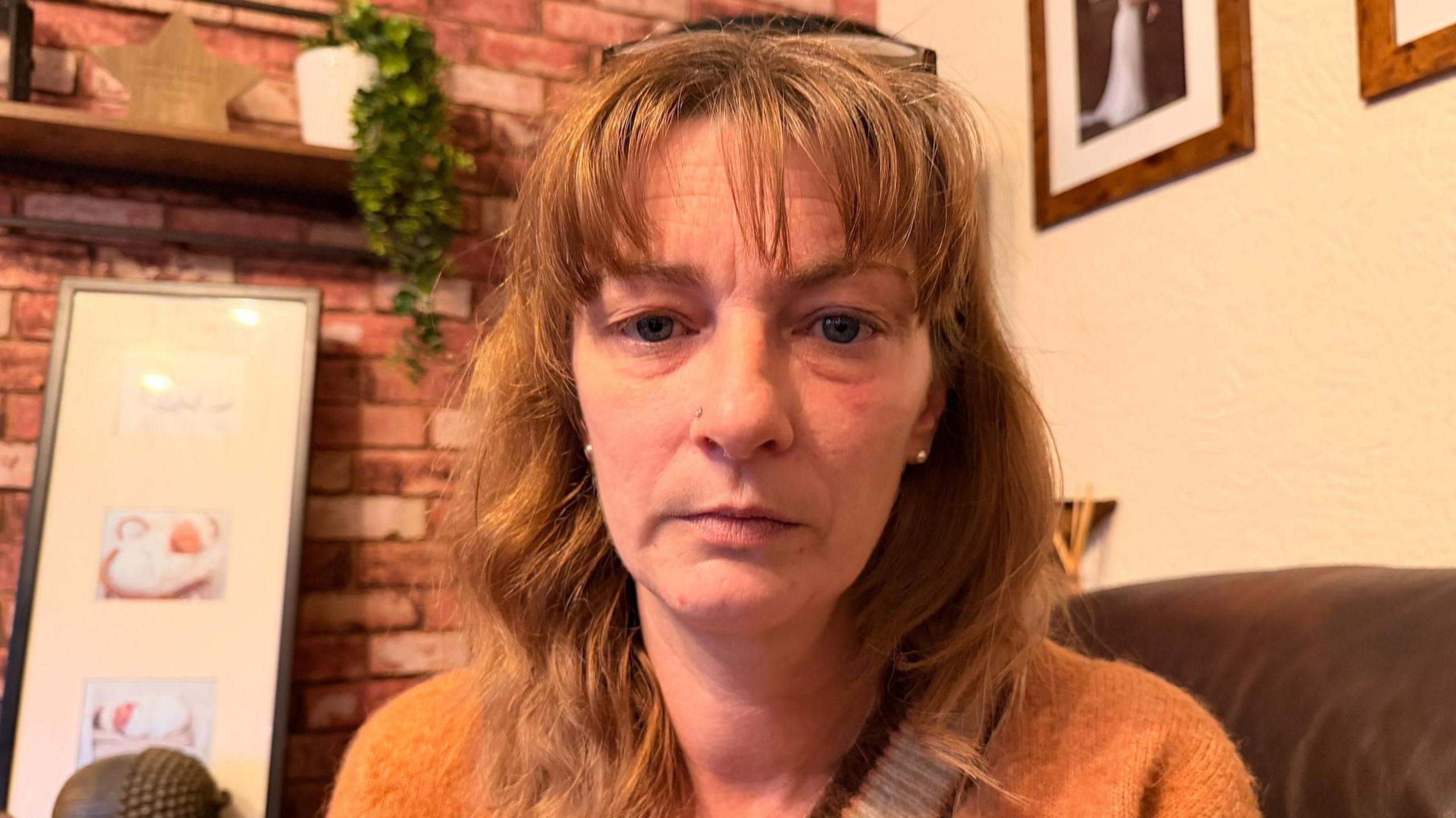 Head and shoulders shot of Helen. She is wearing an orange jumper and is looking directly at the camera. She is sitting on a brown-coloured sofa.
