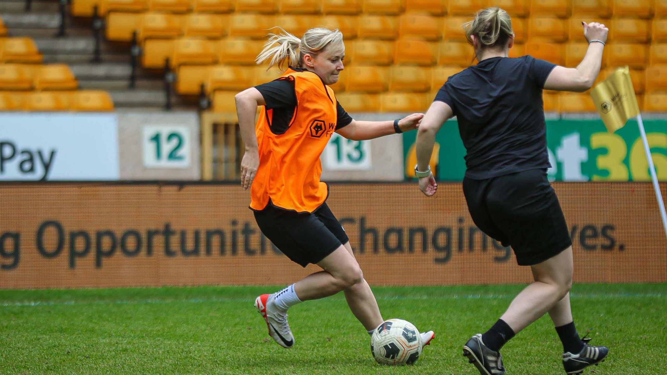 A  woman with blonde hair tied in a ponytail. She is wearing a black t-shirt with an orange bib on it. It has the wolves logo on it and says wolves foundation. She is kicking a football on a pitch. Another woman, dressed in black top and shorts, is running towards her