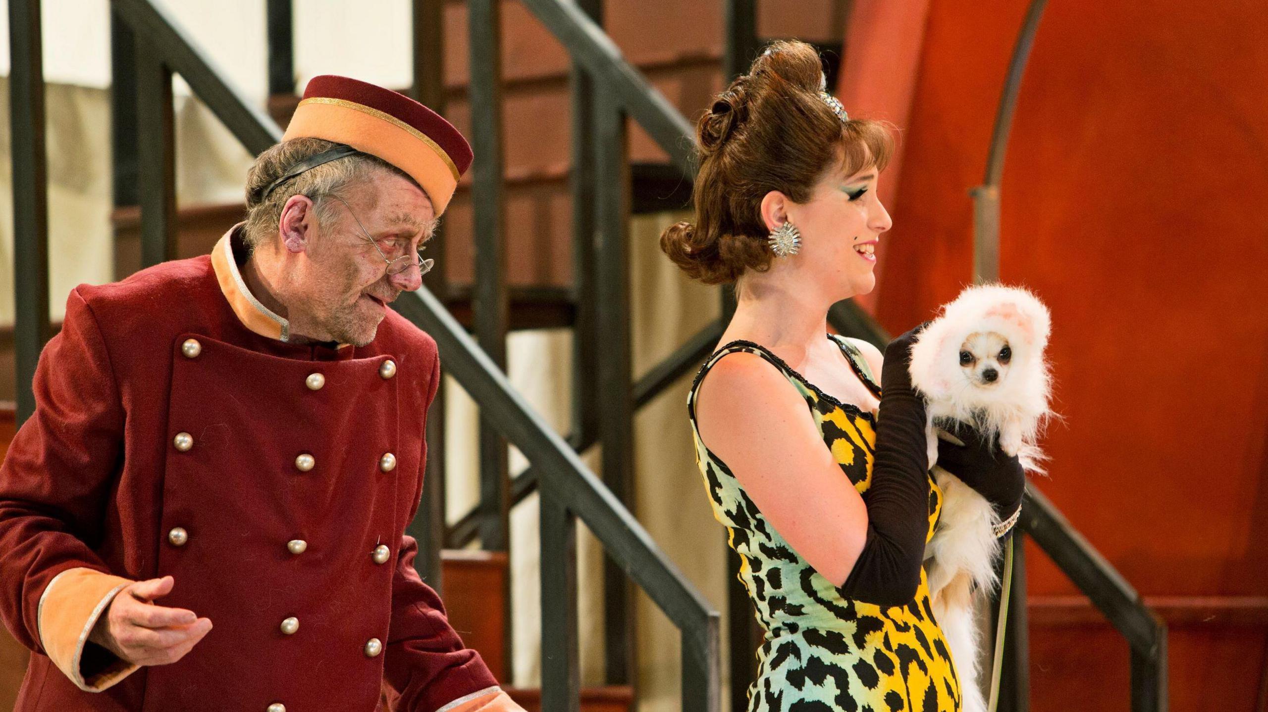 A scene from Scottish Opera's 2014 version of Don Pasquale,  as the character Norina - in a multi-coloured dress, holds Bridget the dog - dressed as a cat, while a porter looks on