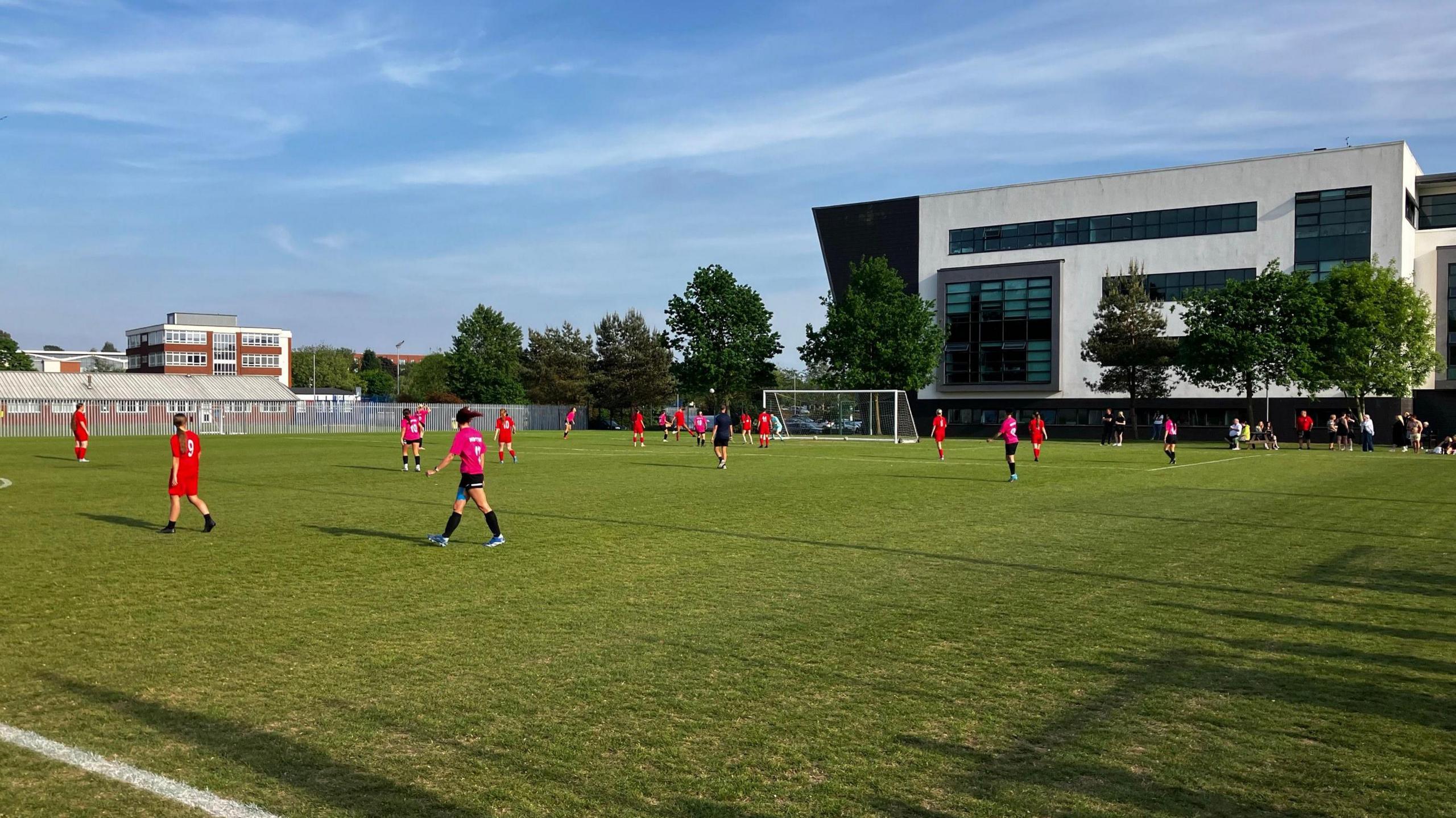 Two women's football teams, wearing red and pink kits, playing a match on an outdoor pitch. A black and white college building is in the background behind some trees.
