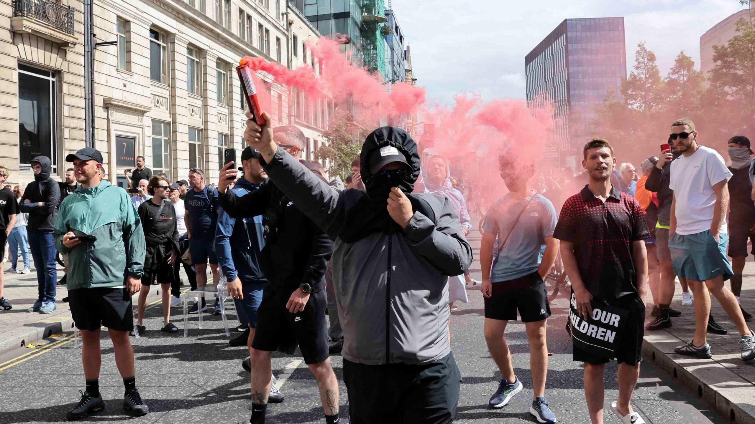 A protesters with his hood up and hiding his face holds up a smoking red flare in Liverpool