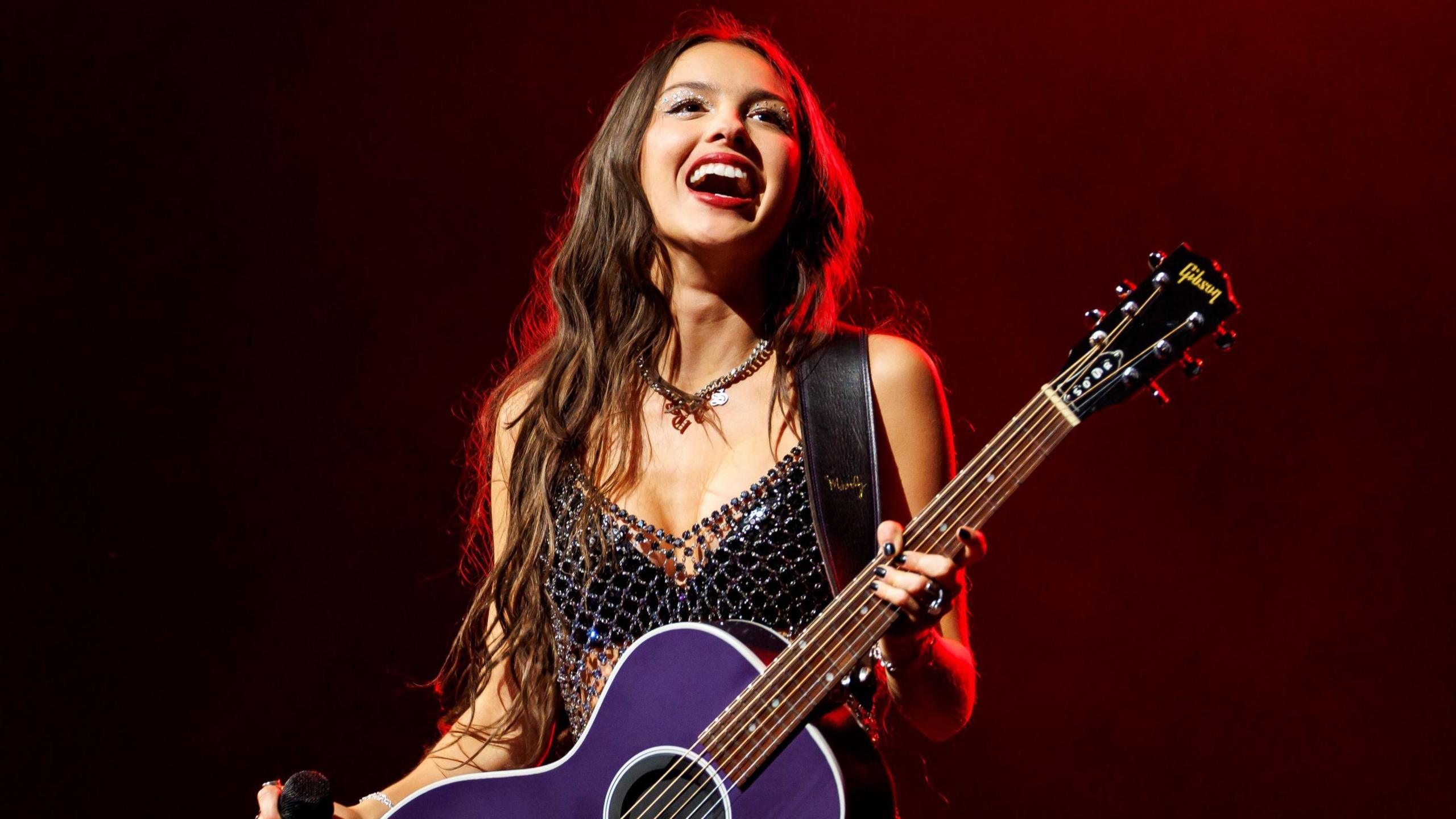 Olivia Rodrigo on stage during her Guts world tour. The singer has long brown hair worn loose and wears a beaded dress over a black bra. She holds a purple guitar and smiles at the crowd. 