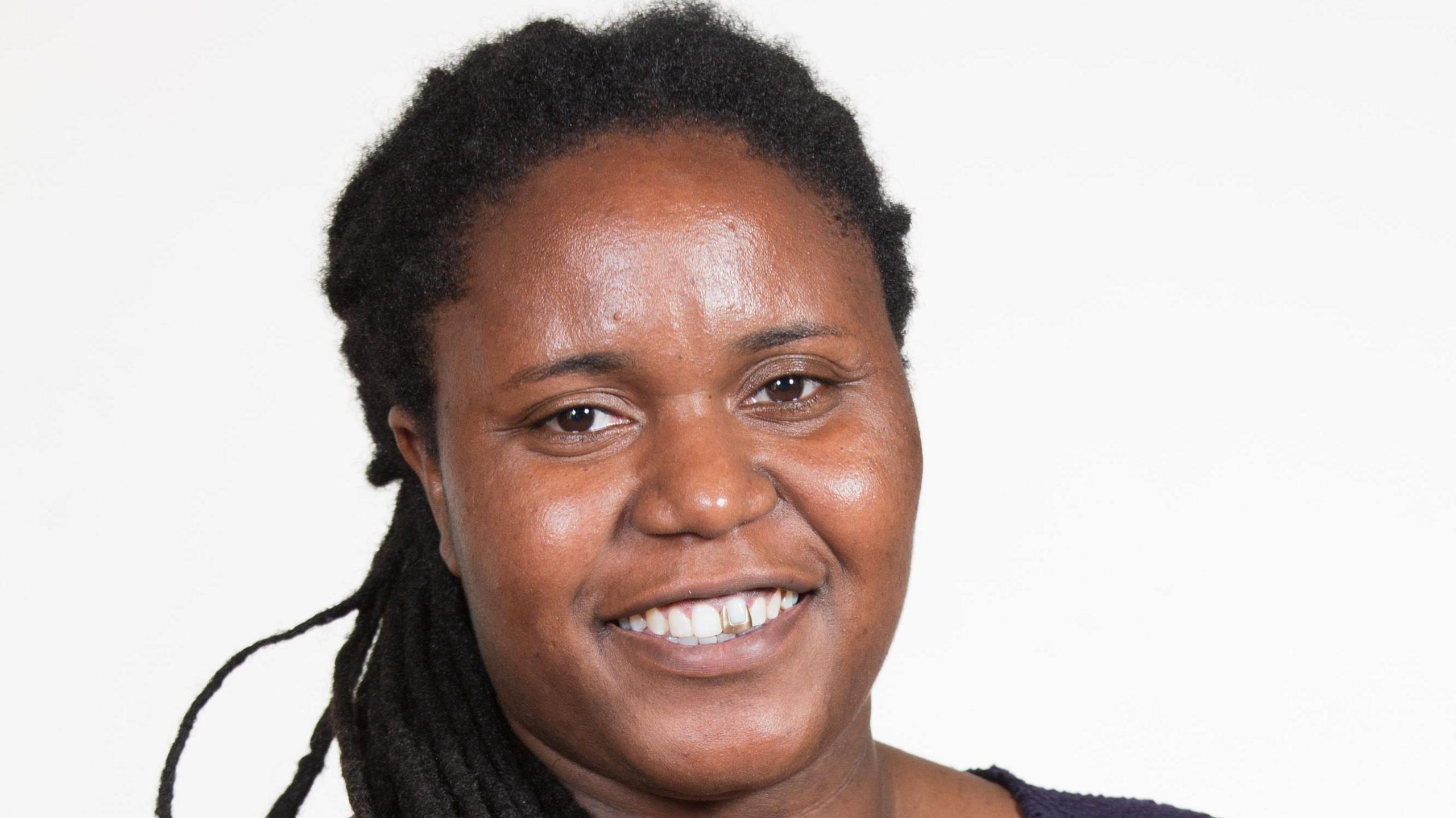 A black woman with long hair in braids smiles at the camera. We only see her face and part of her shoulder which shows she is wearing a dark top.