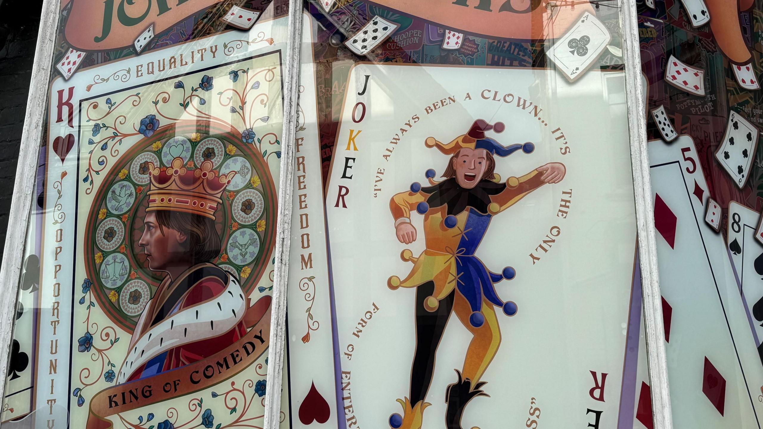 Two playing cards from the mural are displayed in the unit's front window. The one on the left shows Rik Mayall wearing a crown and robes as a king with "King of Comedy" written on a banner underneath him. The one on the right shows the comedian as a joker in a blue and yellow jester outfit. 