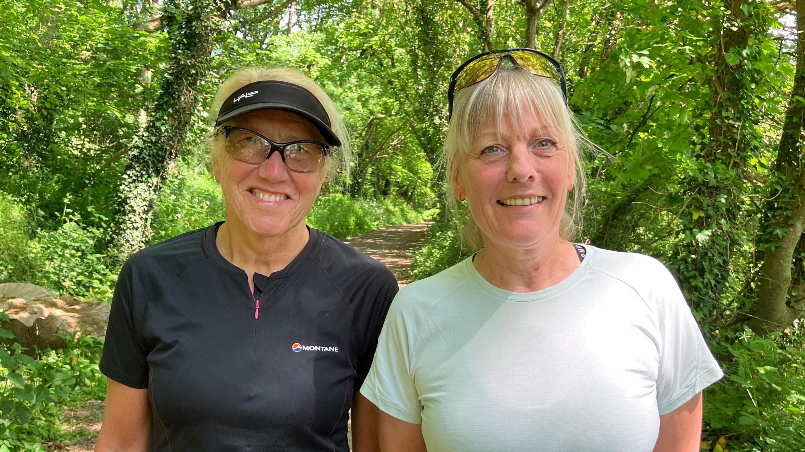 Two women in training clothing in a woodland area. The one on the left has a black top, glasses and a sun visor. The woman on the right , has a pale top, and yellow lensed sunglasses on her head.