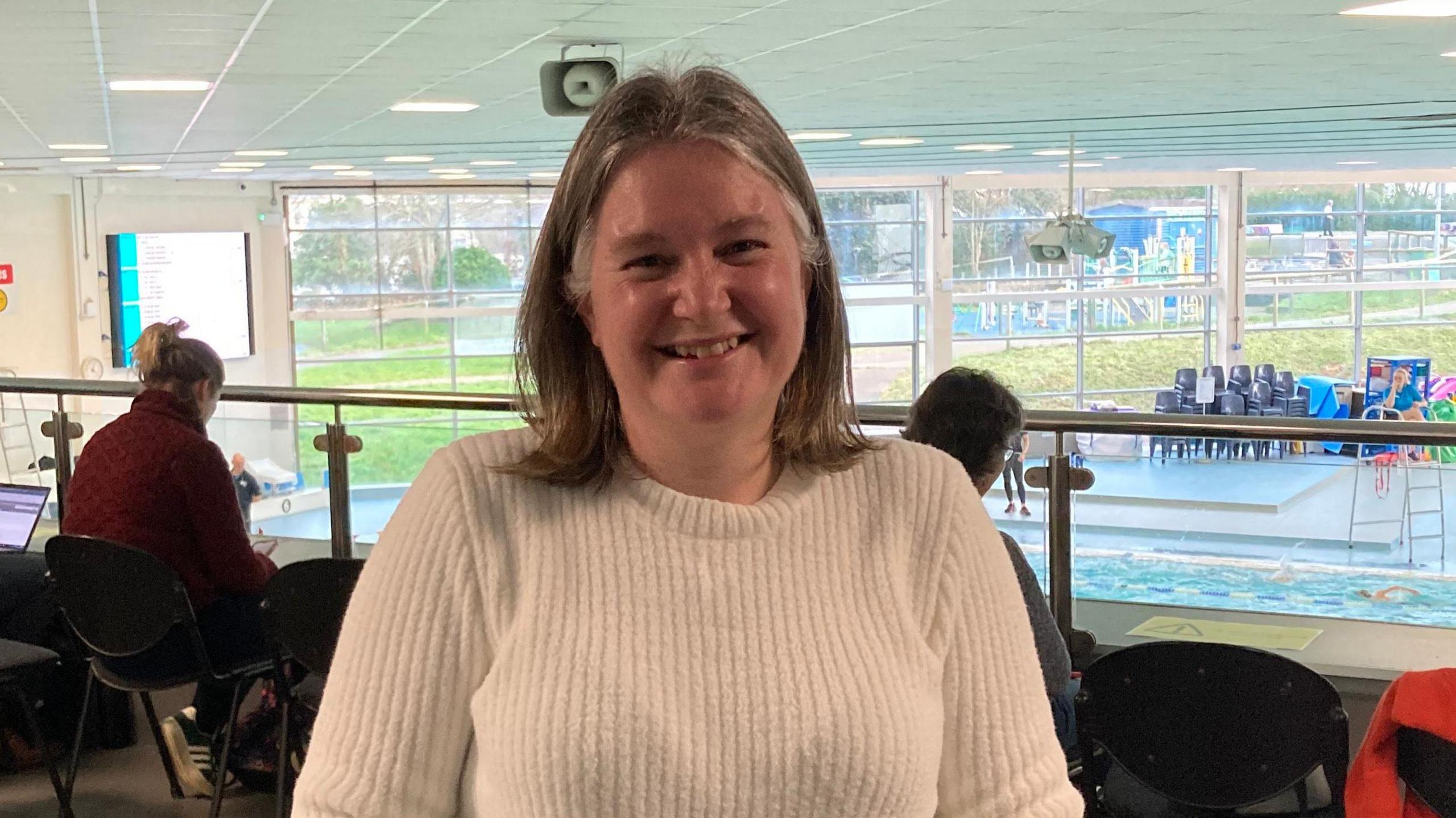 A white lady wearing a white jumper with a brown bob, smiles at the camera in front of a swimming pool.