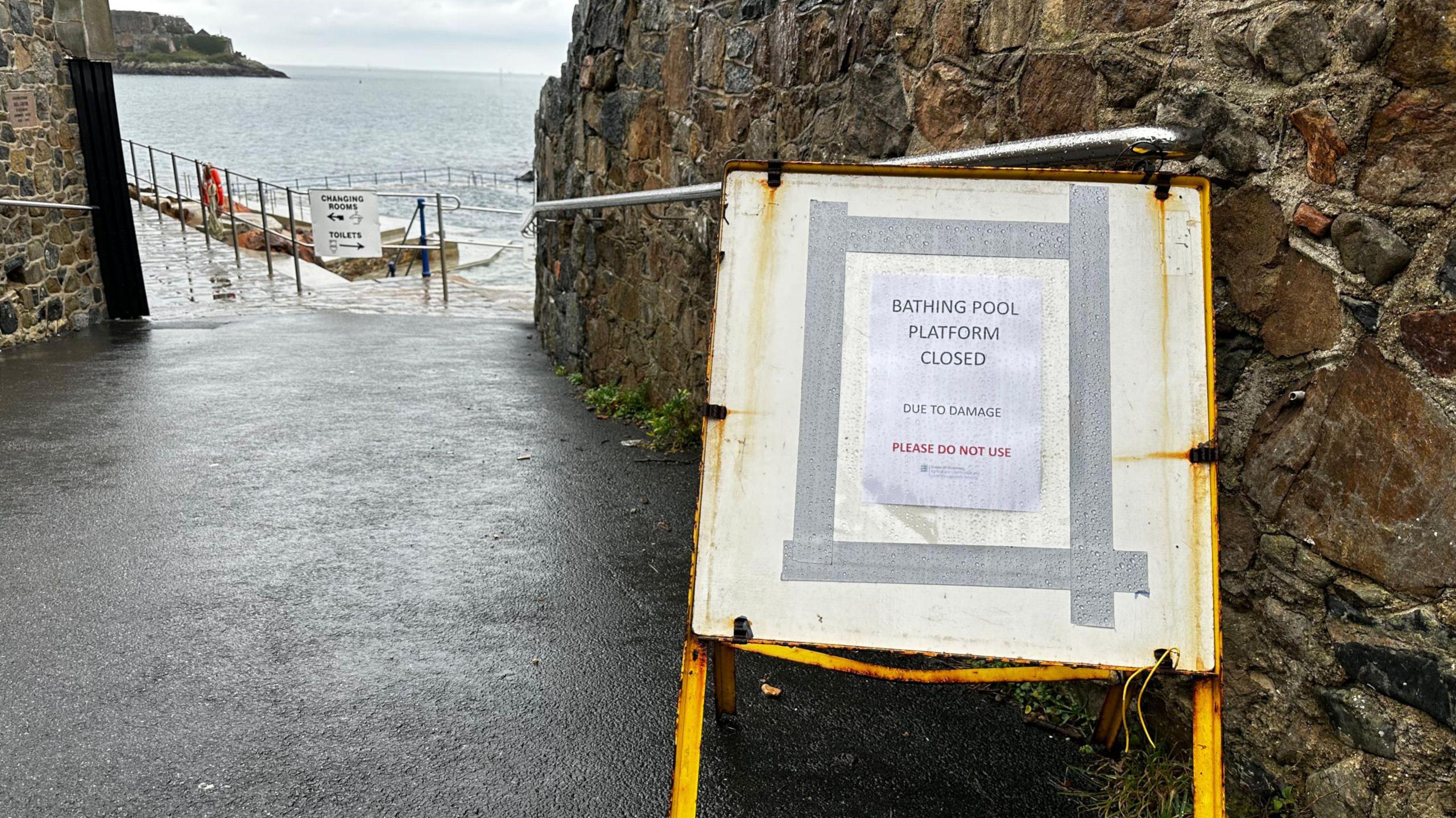 A sign saying the diving platform is closed on a slope leading down to an outdoor pool with walls and handrails on either side. Beyond the pool is the sea and part of Castle Cornet.