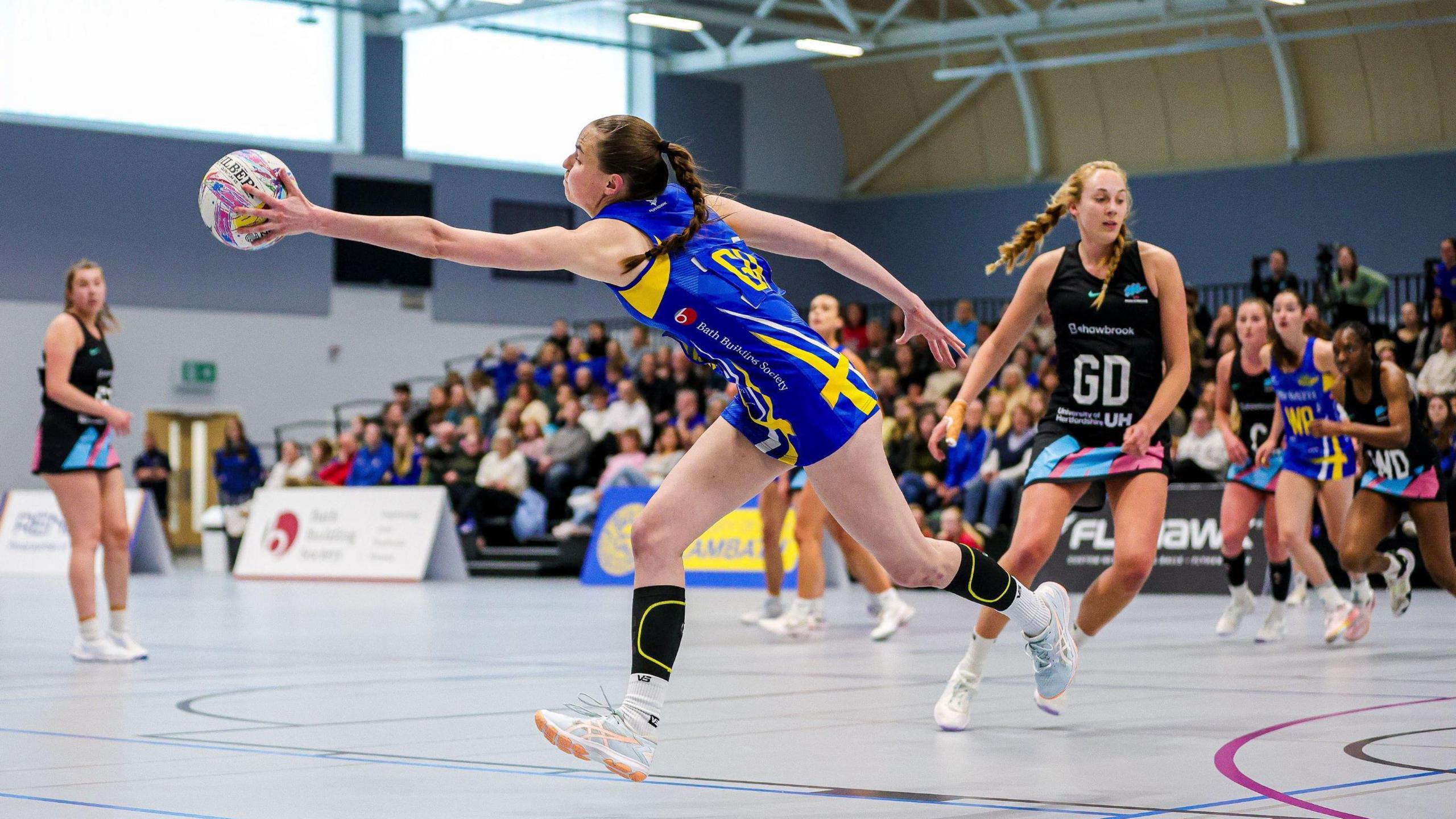 A player from Team Bath stretches out to catch the ball during a netball match against the London Mavericks. She is wearing a blue and yellow kit and in the background other players from each side are watching on, with the London players wearing a black kit with pink and blue edges.