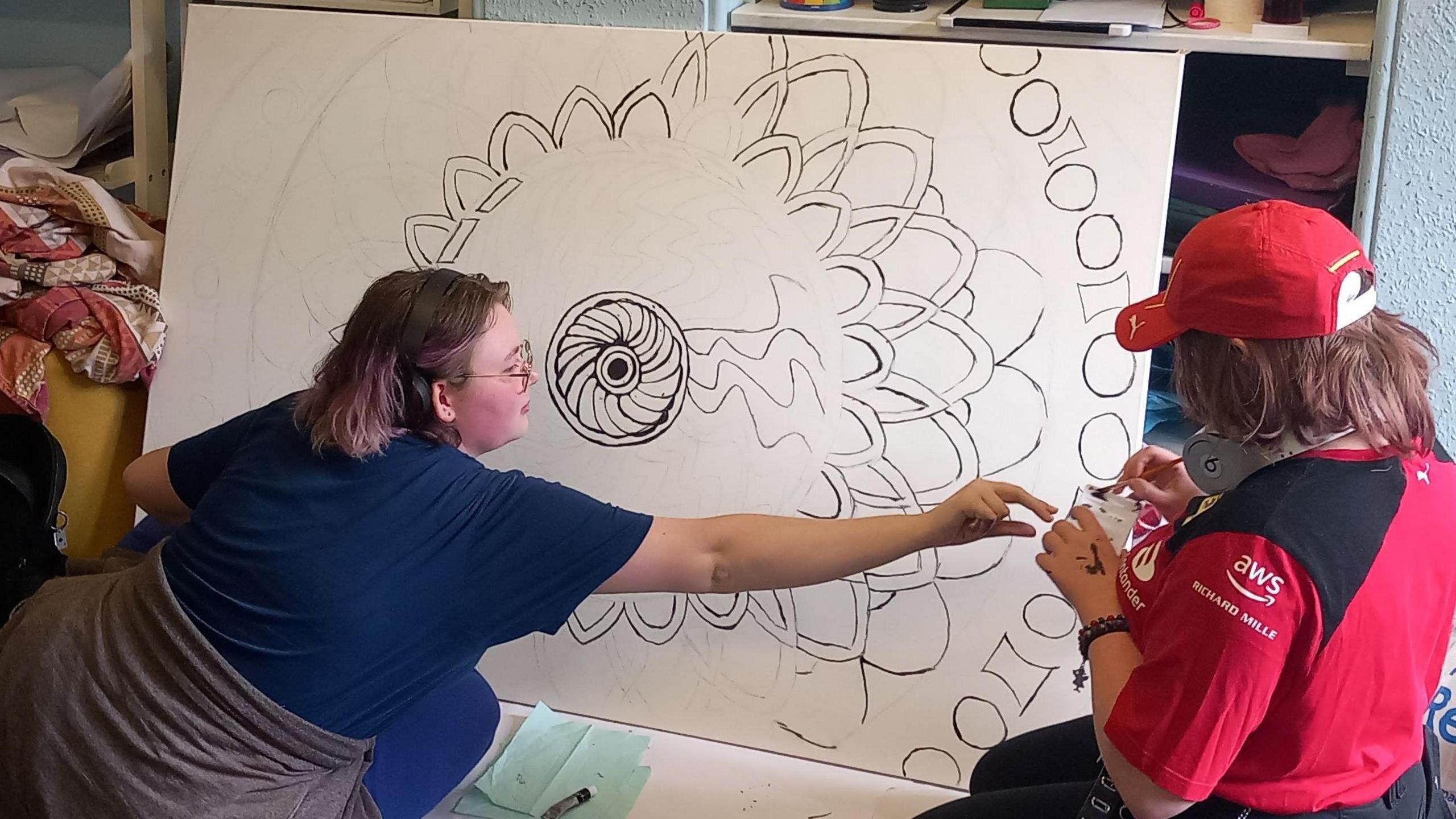 Two young women in front of a large white canvas with a flower design starting to emerge in black ink