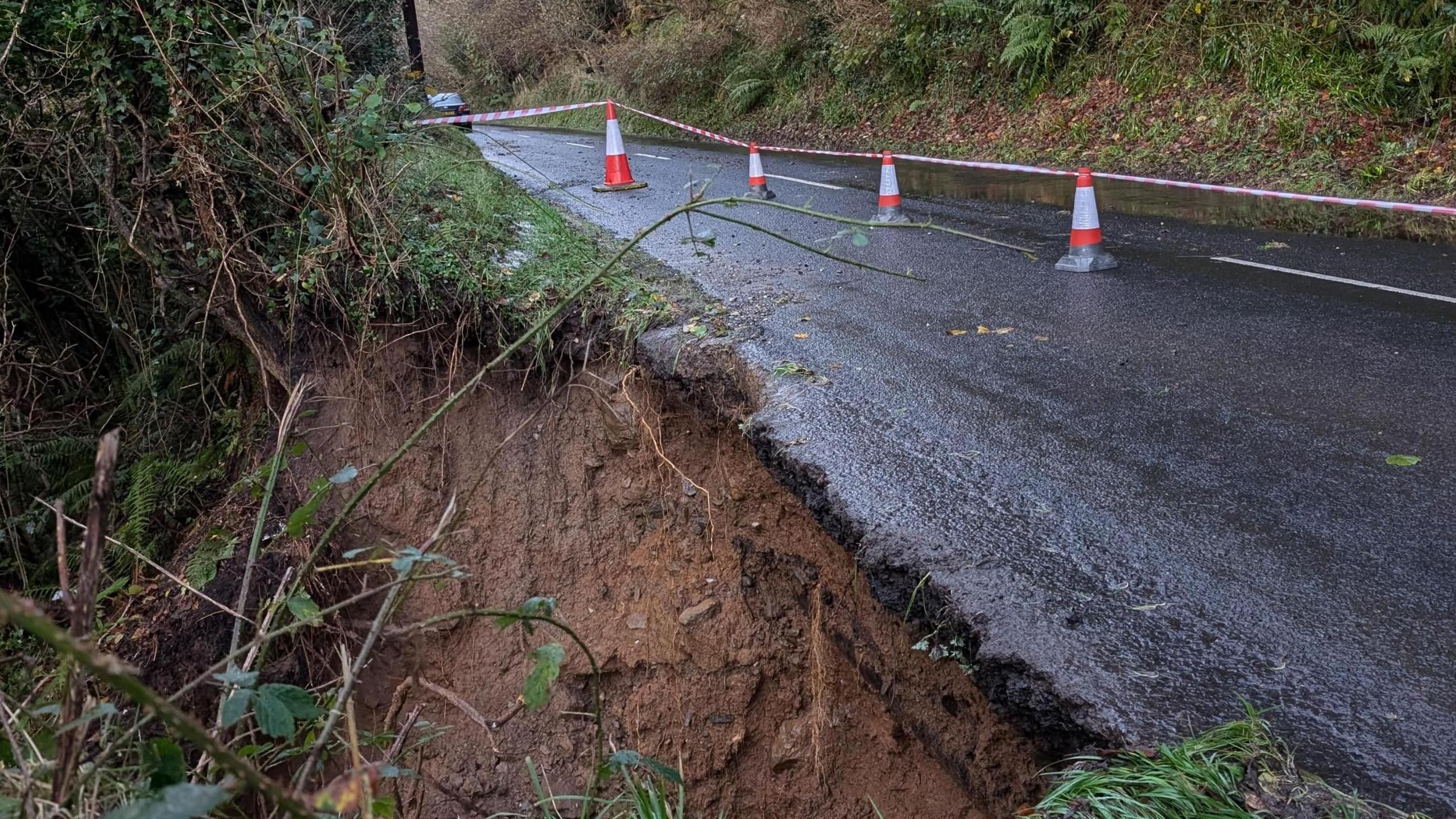 A landslide on the Glenshesk Road following Storm Bert.  A section of the roadside verge has fallen down a hill.  The section of road has been cordoned off with traffic cones and plastic tape.  