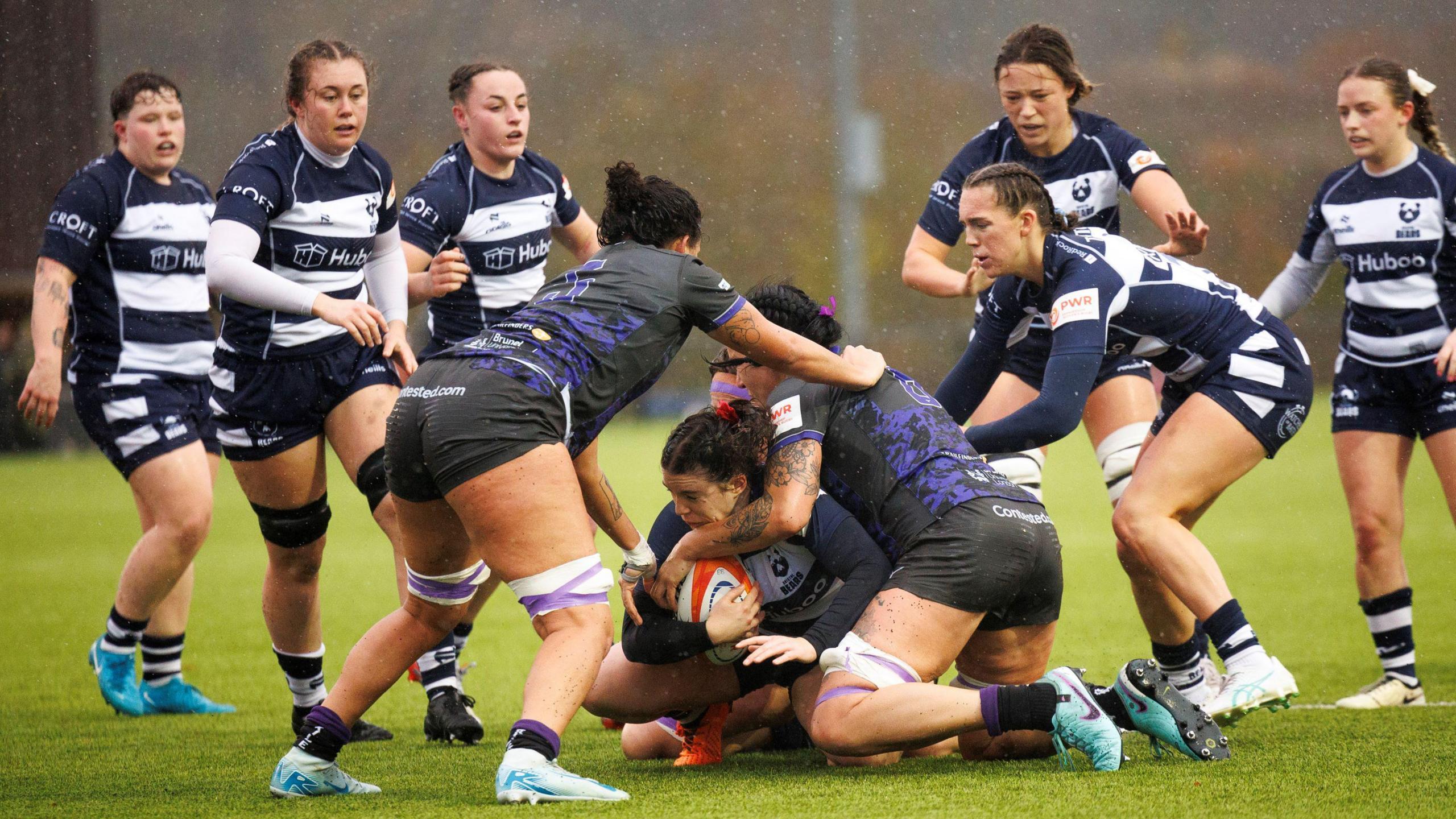 Players from Bristol Bears Women and Ealing Trailfinders contest the ball during their Allianz Premier 15s match in Bristol. The weather is wet with rain falling. The Bristol players are in their home kit of thick blue and white hoops, while the Ealing players have a black kit with a purple patter