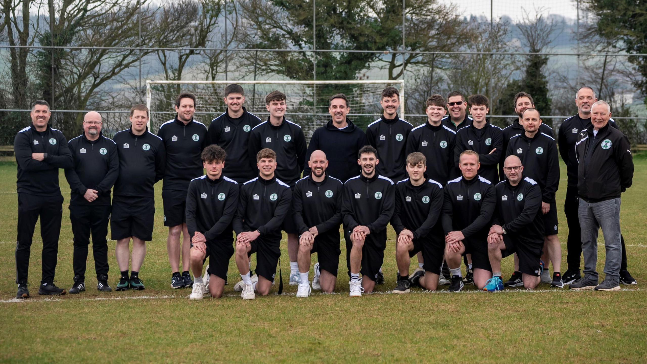 Players from Somerset football club Tunley Athletic FC line up in a team shot for the camera along with former Manchester United player Gary Neville. The players are in an all-black kit