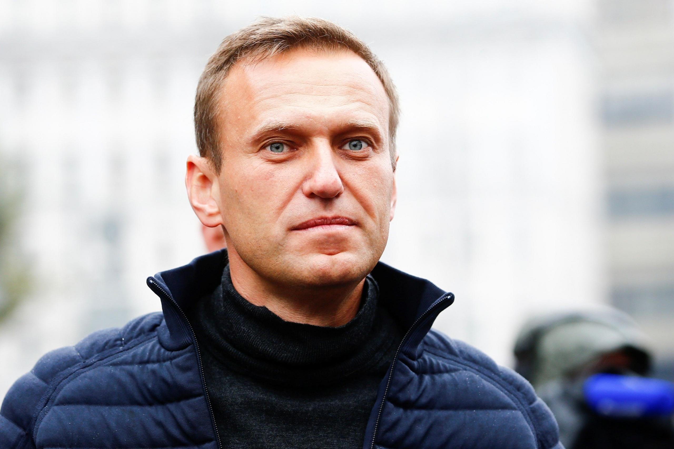 Alexei Navalny has light hair and clear blue eyes in this photo and appears to be staring boldly at something beyond the camera. The photo is taken outside on what appears to be a cold morning. He has a dark sweater on under a blue puffa jacket, unzipped.