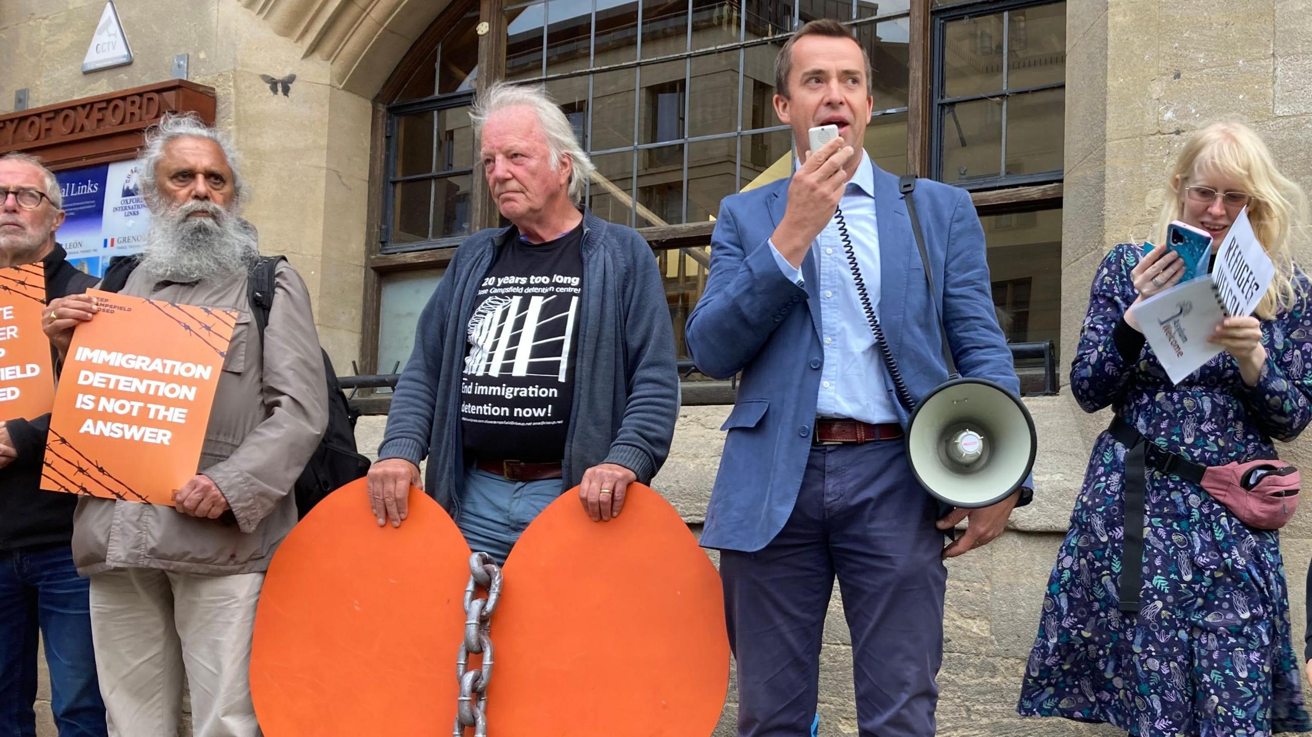 MP Calum Miller stands right of centre, wearing a blue suit jacket and holding a megaphone, on his right is a woman in a patterned dress reading her phone and holding a pamphlet, and on his left are two older men, one holding a large orange heart with a chain across it, and the other has a sign saying "Immigration Detention is Not The Answer"