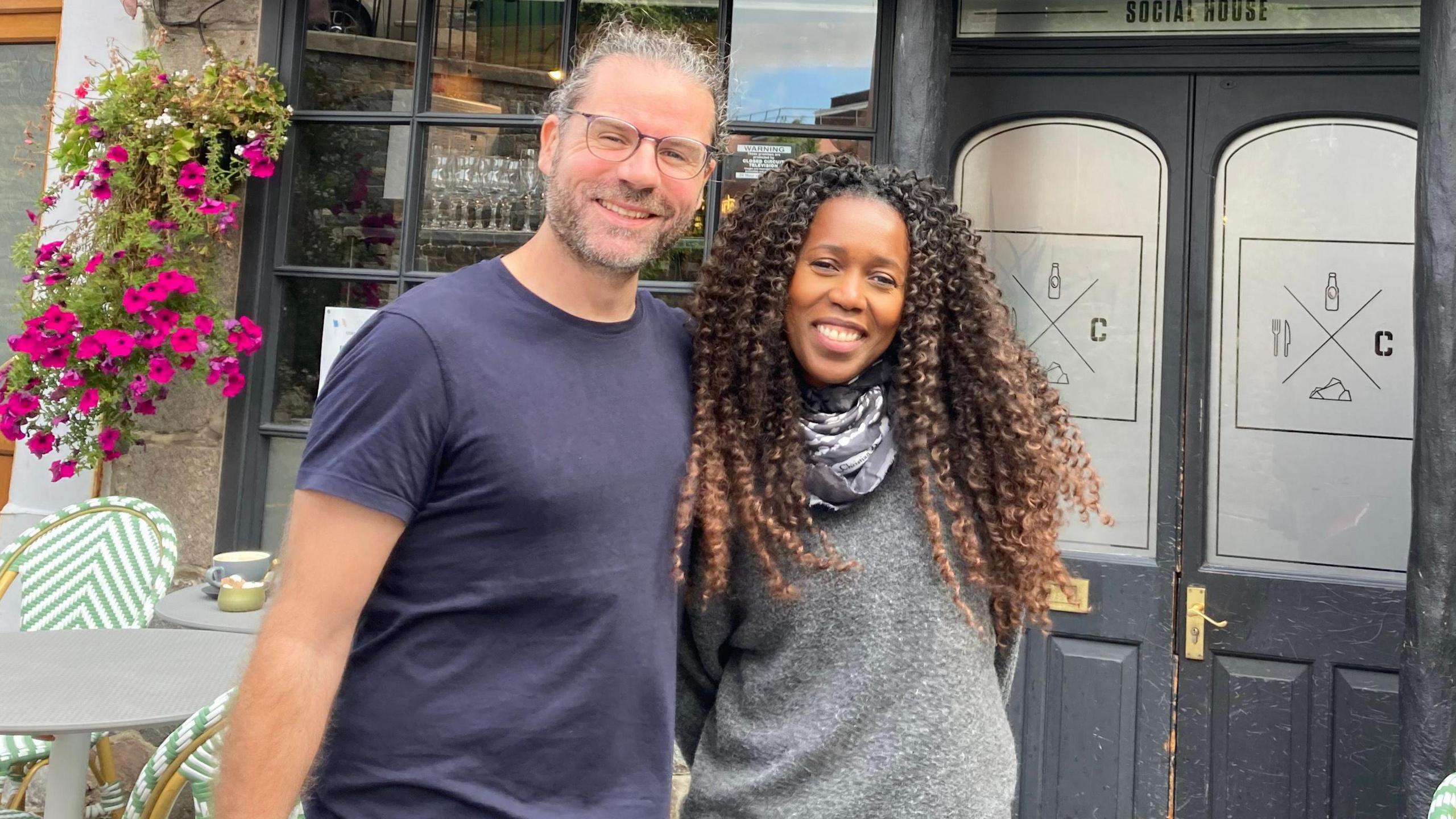 A man standing to the left in a blue t-shirt and glasses next to a black woman with long curly hair. They are standing in front of a building with a black door.