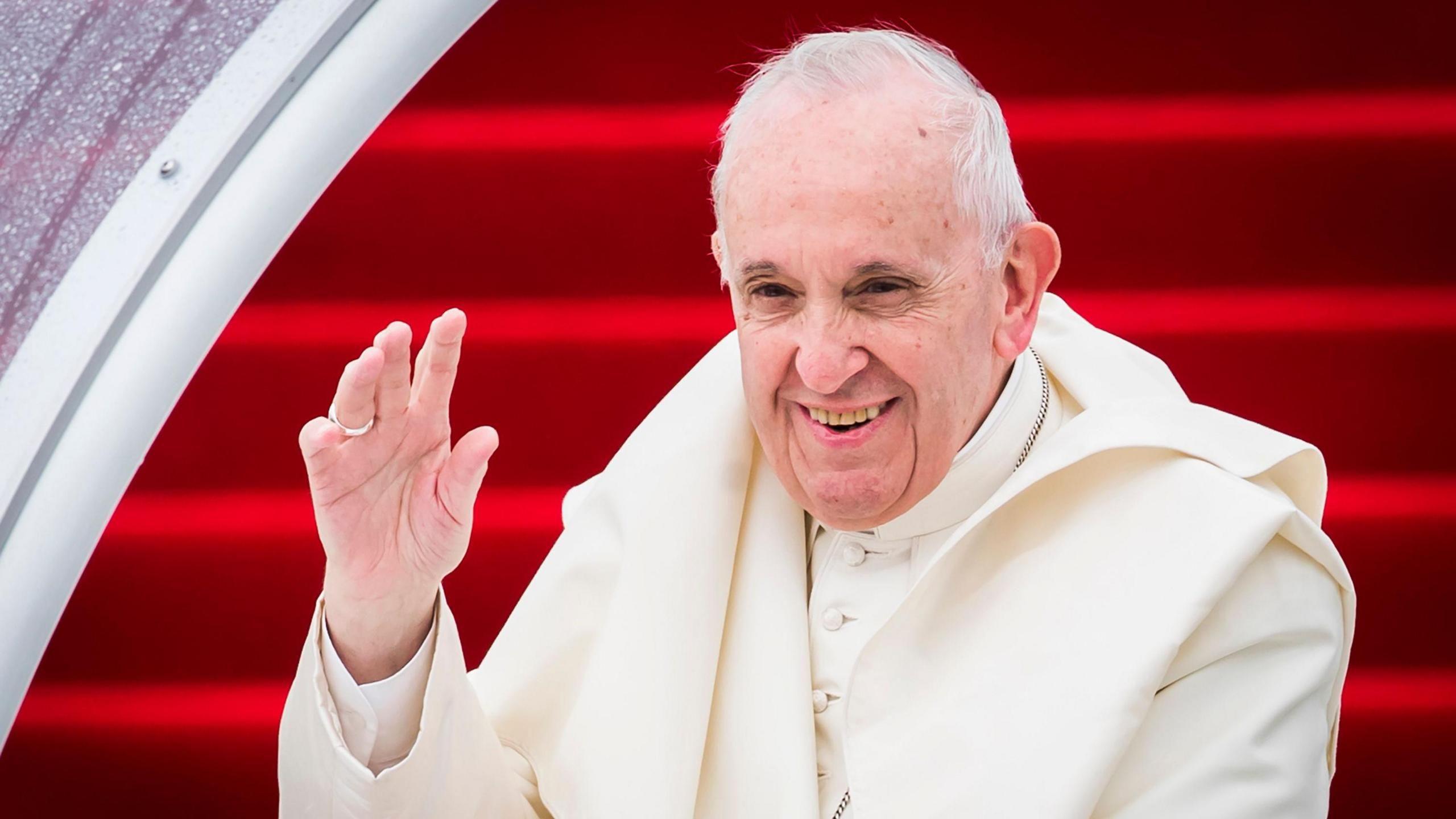 Pope Francis wearing white robes waving to someone off camera.