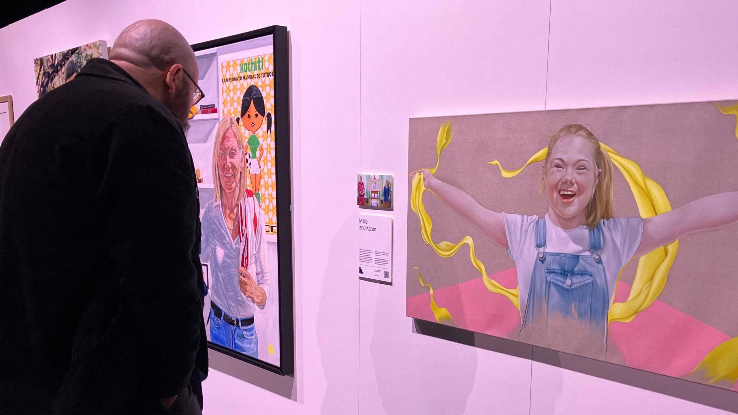 A bald man with a beard reads a plaque next to a painting of a young girl smiling with her arms outstretched on a gallery wall. 