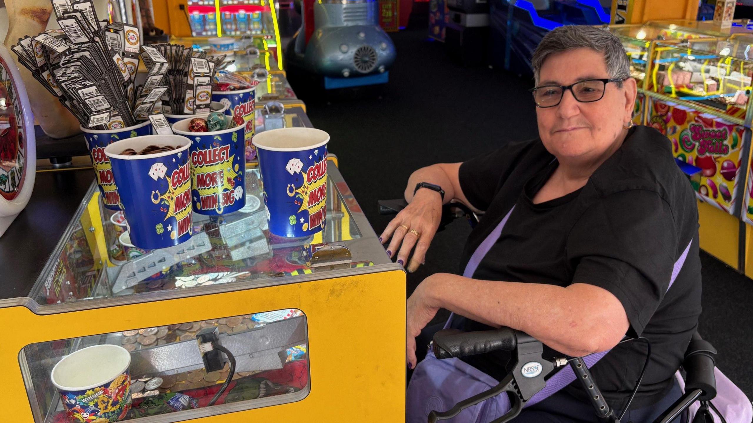 Chris Dady sits next to a 2p arcade machine. On the top of the machine are paper cups containing hundreds of tickets which can be exchanged for small prizes