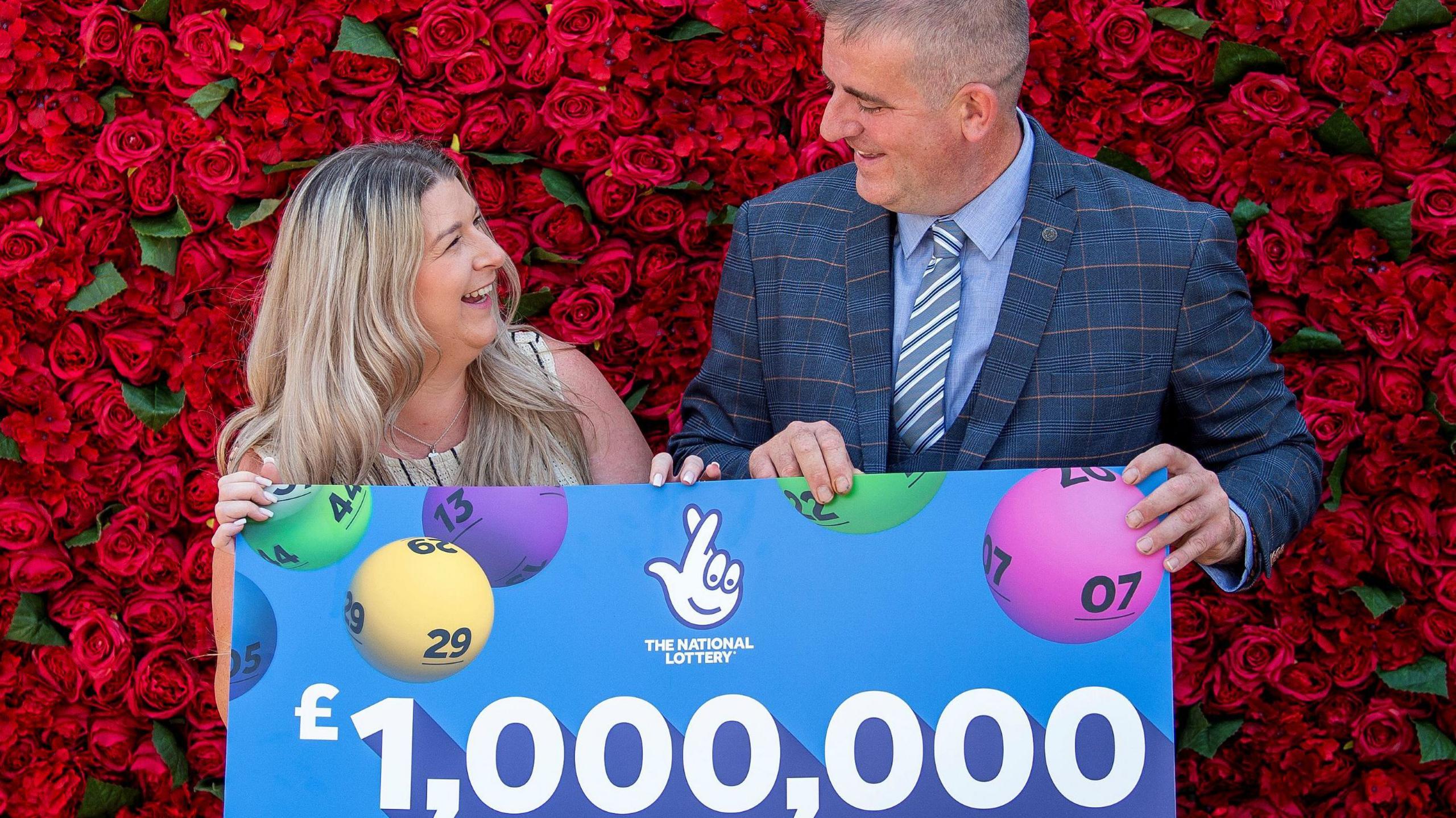 Darren McGuire, who has short greying hair, in a blue check patterned suit with sky blue shirt and blue and white club tie holds a giant cheque for £1m with partner Laura who is wearing khaki sleeveless top