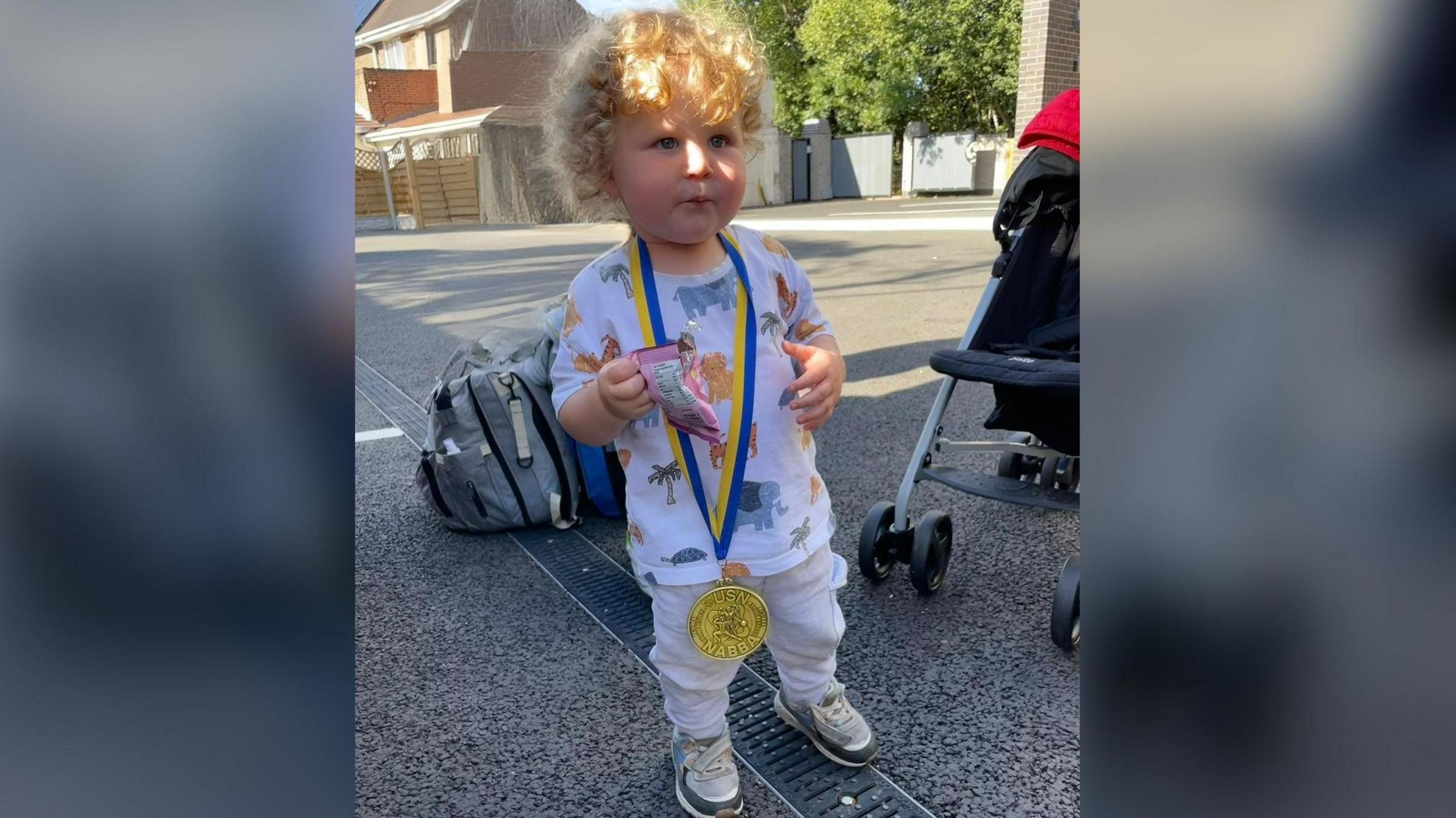 Hunter is standing and holding a packet of sweets. He has short curly blonde hair. He has a gold medal with a yellow and blue ribbon strap around his neck.