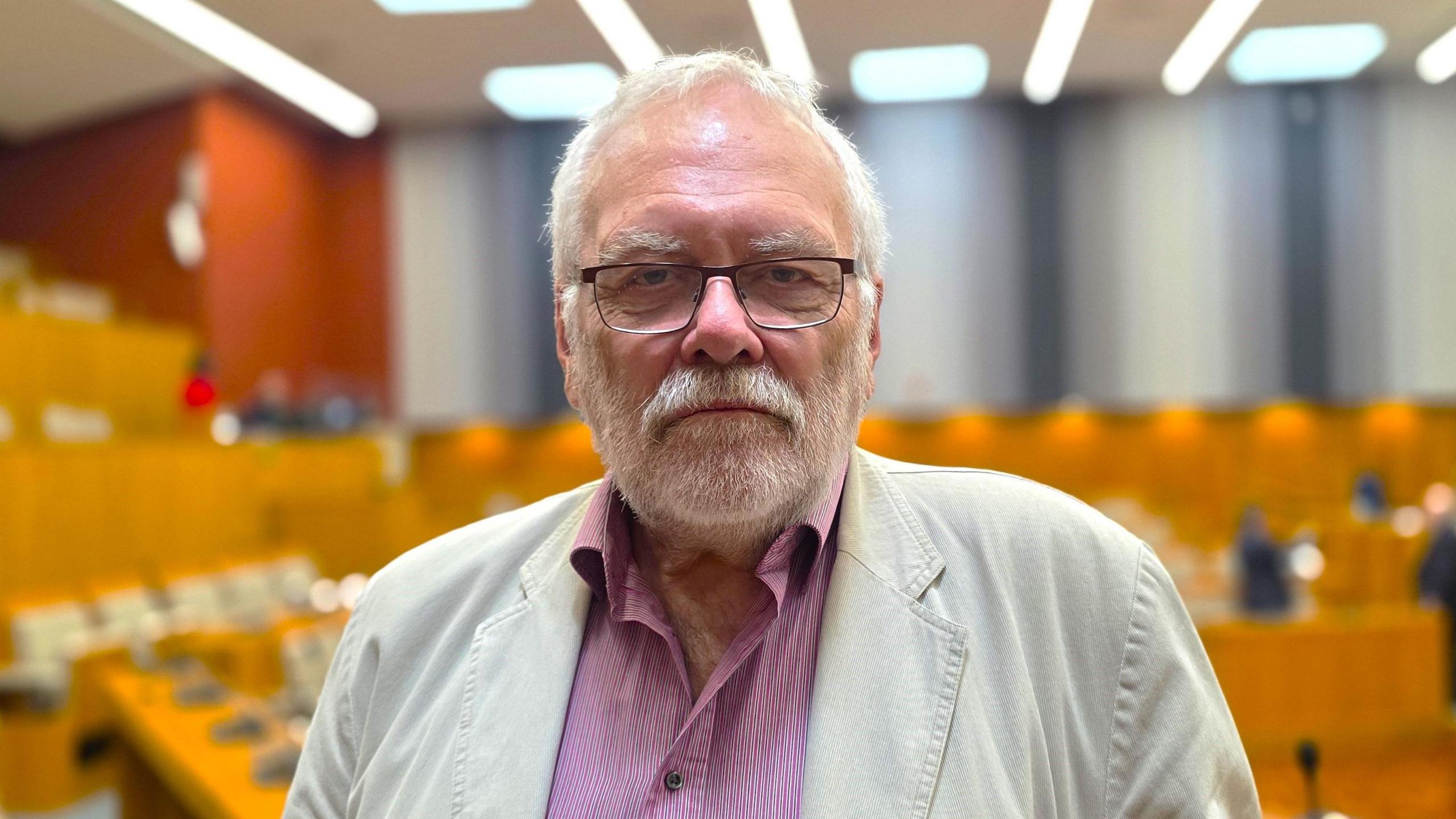 Steve Morphew is wearing a light, cream linen jacket over a red and white pin-striped shirt. Behind him, we can see an (out of focus) council chamber.