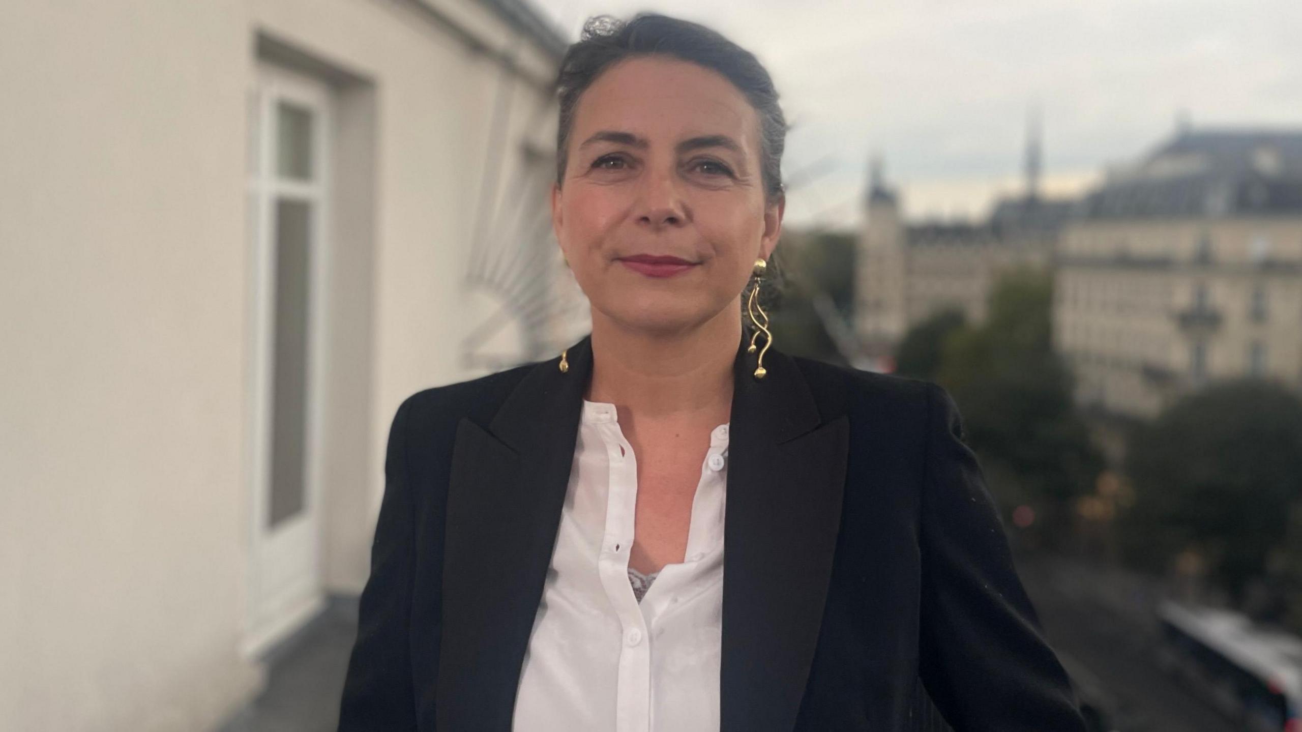 Elodie Tuaillon-Hibon is photographed on a balcony in Paris. She is looking straight into the camera and wearing a white shirt and black jacket with large gold earrings.