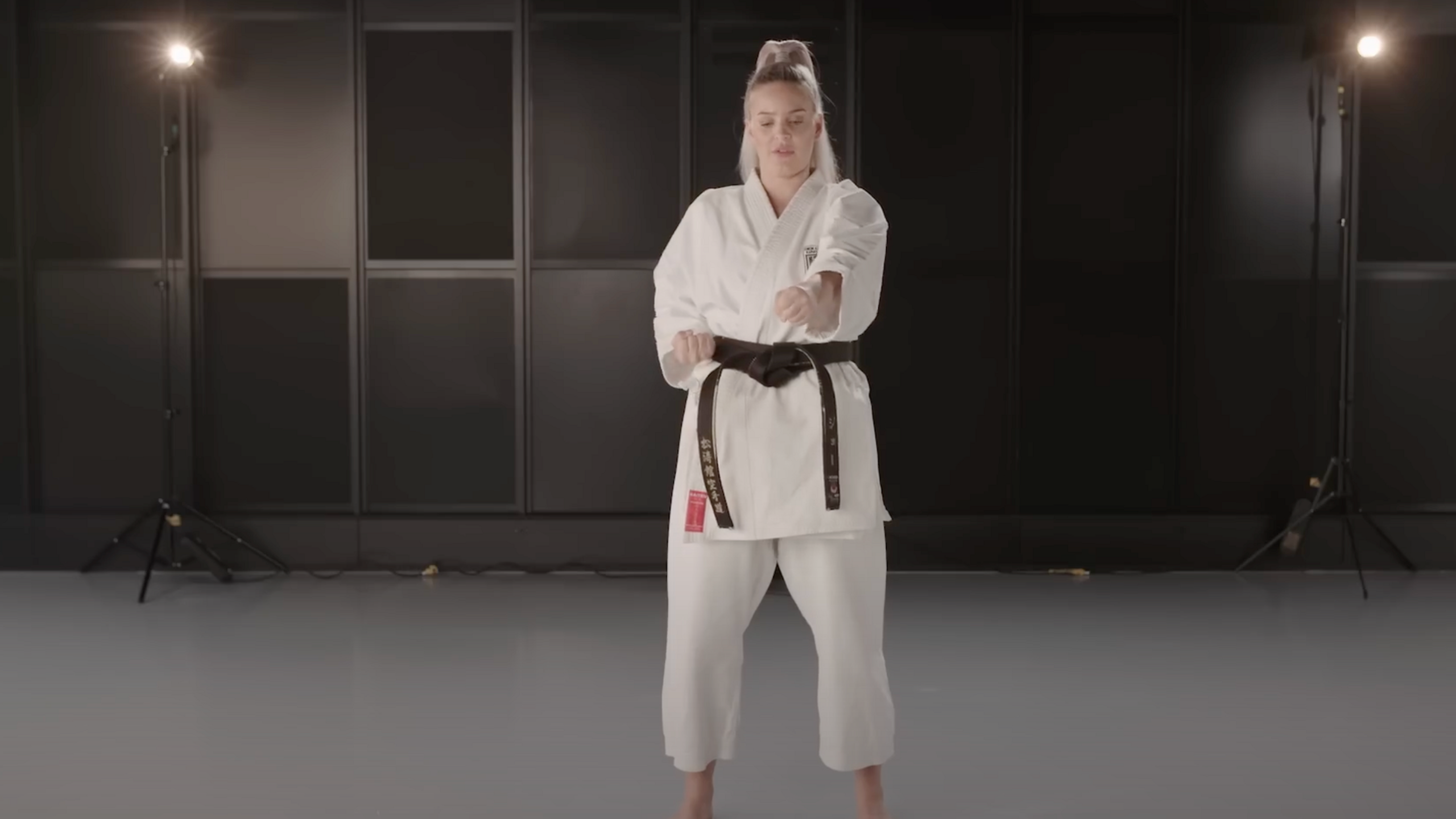 Anne-Marie in a white karate outfit or Gi with a black belt demonstrating a punch. 