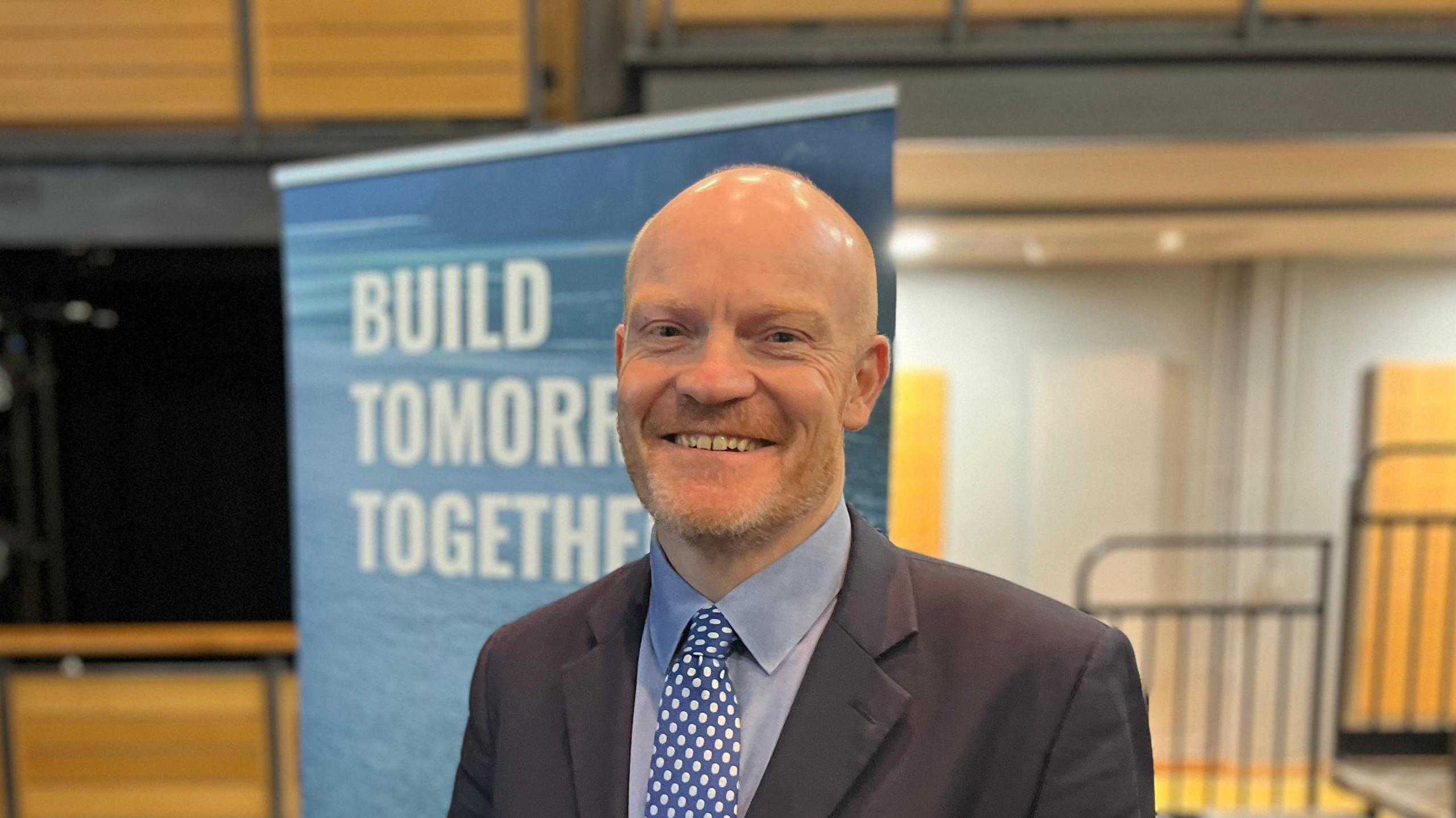 Gavin St Pier pictured wearing a dark coloured suit jacket, a grey-ish shirt and a blue and white polka-dotted tie. Mr St Pier has stubble and a bald head.