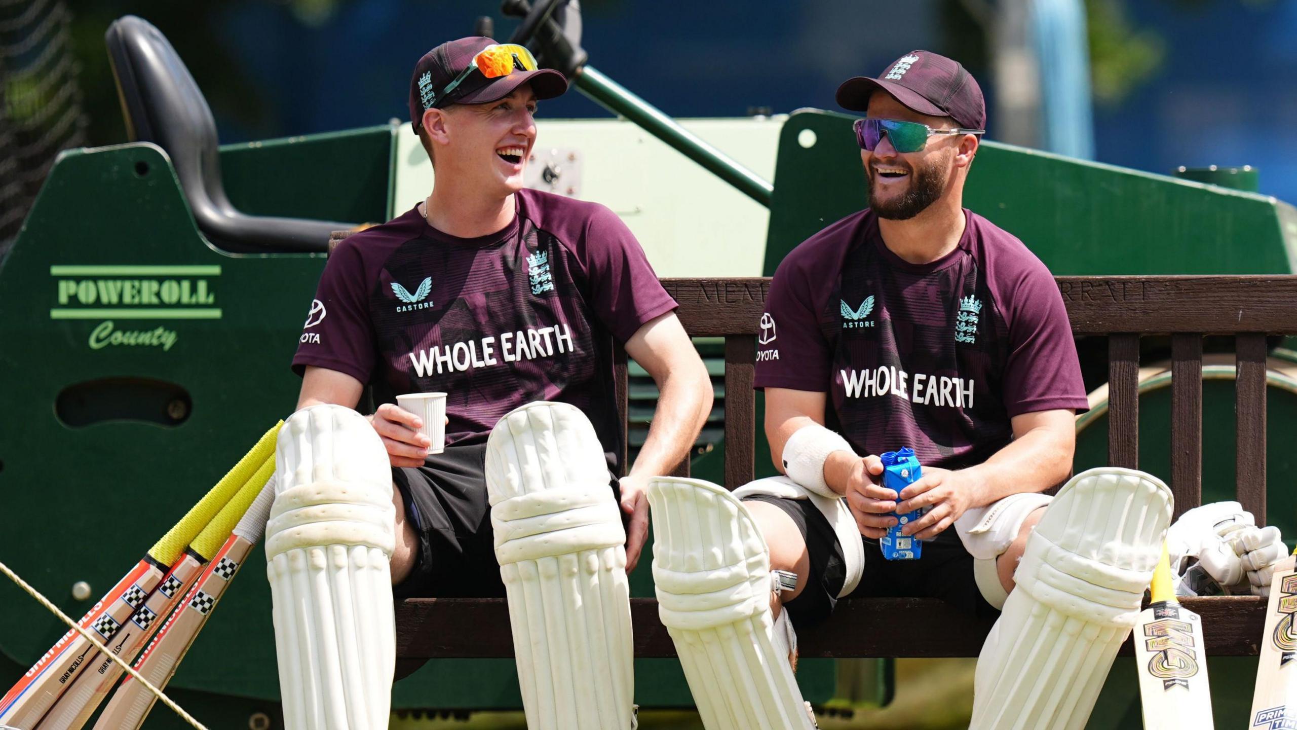 Harry Brook, left, and Ben Duckett share a joke at training. They are sat on a bench in front of a tractor. Three bats are resting against Brook and two beside Duckett