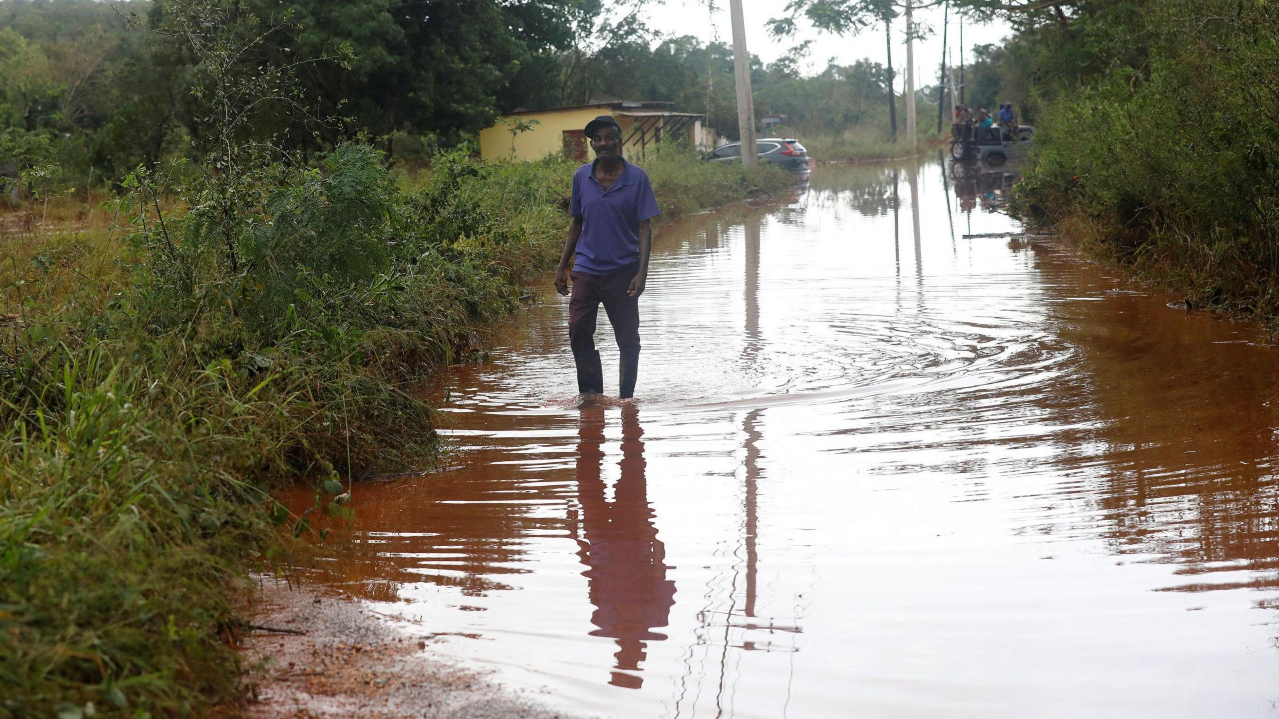 A man in a purple top and dark trousers stands in the middle of a road submerged in brown muddy water. 