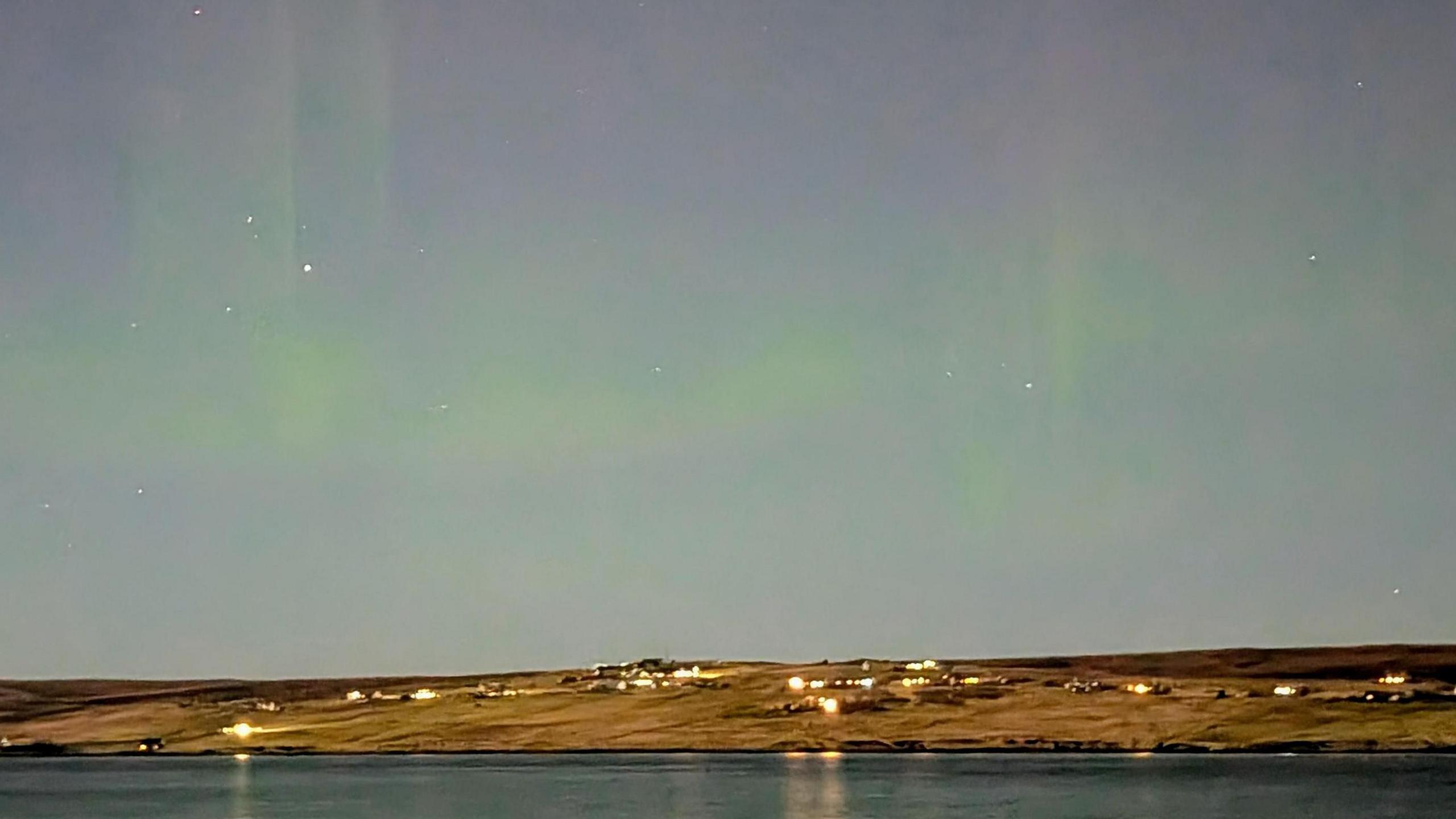 A hint of green aurora in the evening sky over water and houses in a rural area