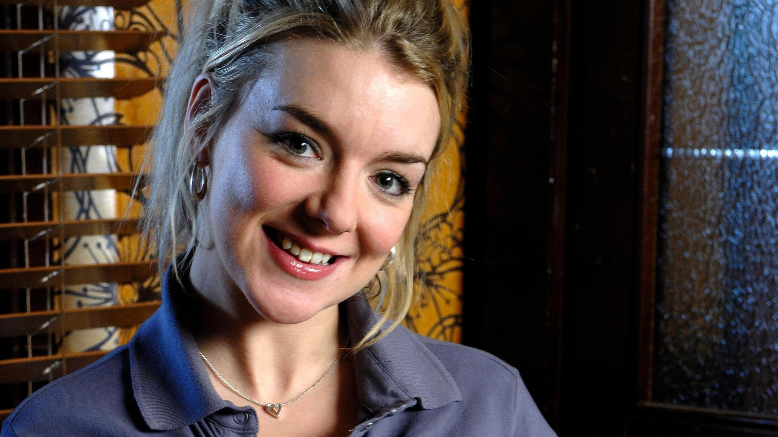 Sheridan Smith. She has blonde hair and smiles as she looks into the camera. She is standing in front of a wooden blind and a wall covered in amber and black wallpaper. She is wearing a blue polo shirt, silver hoop earrings and a silver pendant necklace. 