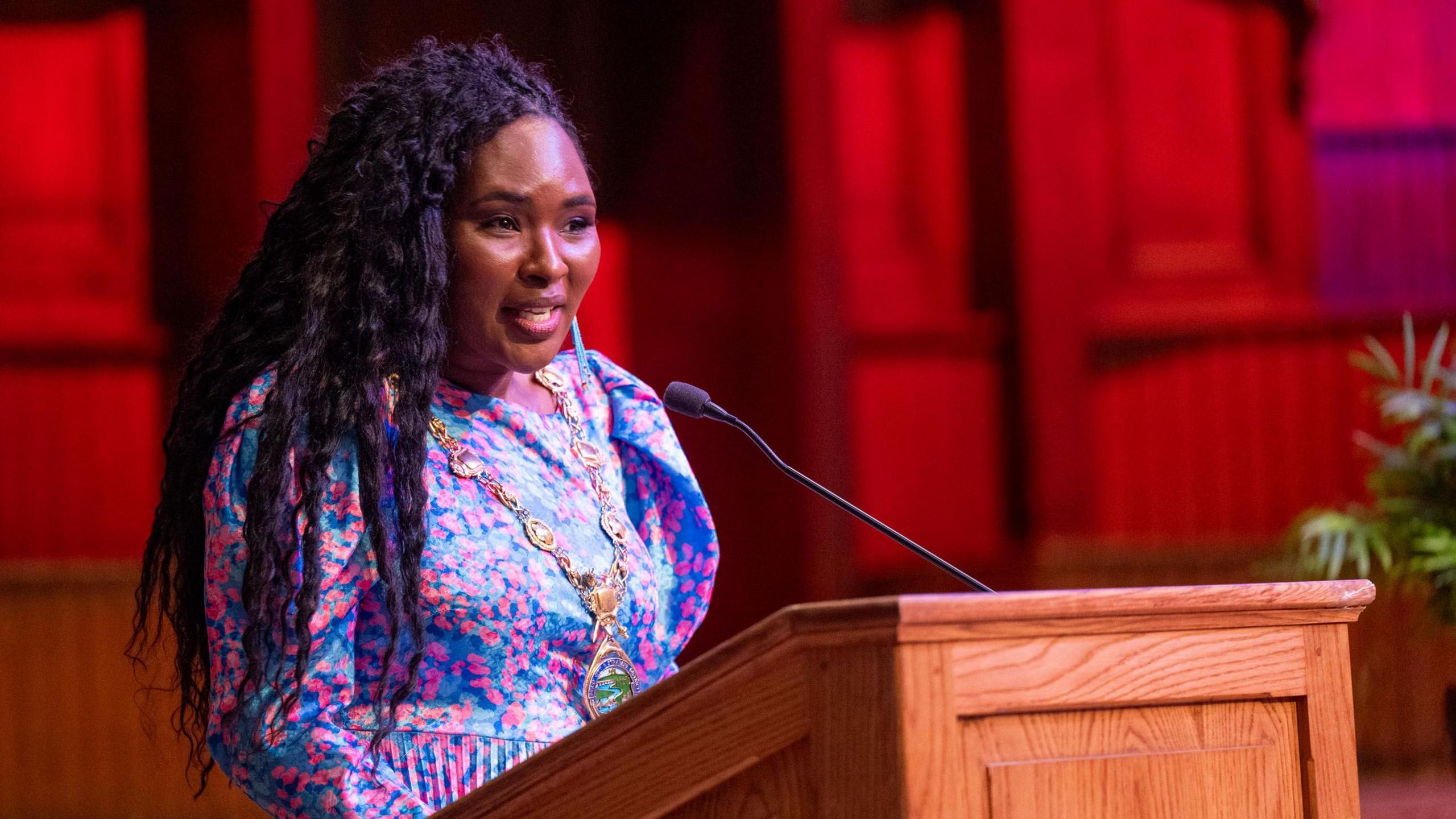 Lilian is speaking at the pulpit. She has black long wavy hair and is wearing a blue dress with pink love hearts on it. 