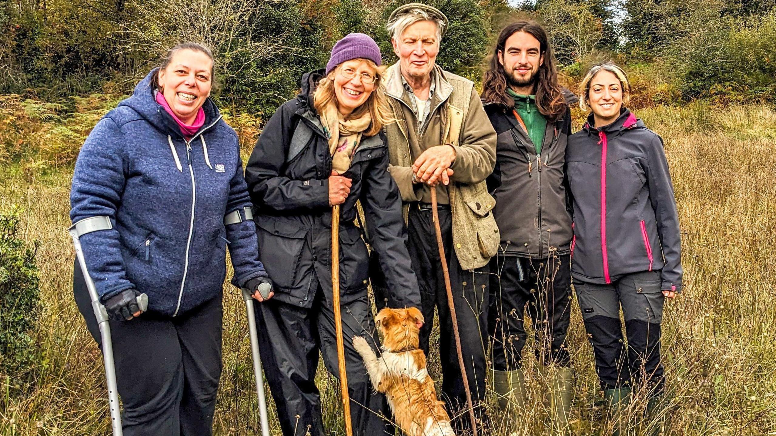 Five volunteers stand together in a field. They're looking into the camera. A small orange dog is standing up on a woman's knees and she's petting him. There is grass and bushes surrounding them.