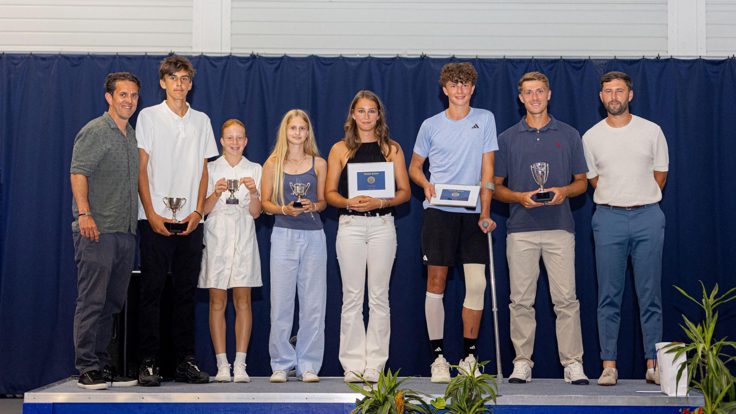 Eight people are standing in a row on a stage holding up trophies. They are all smiling.