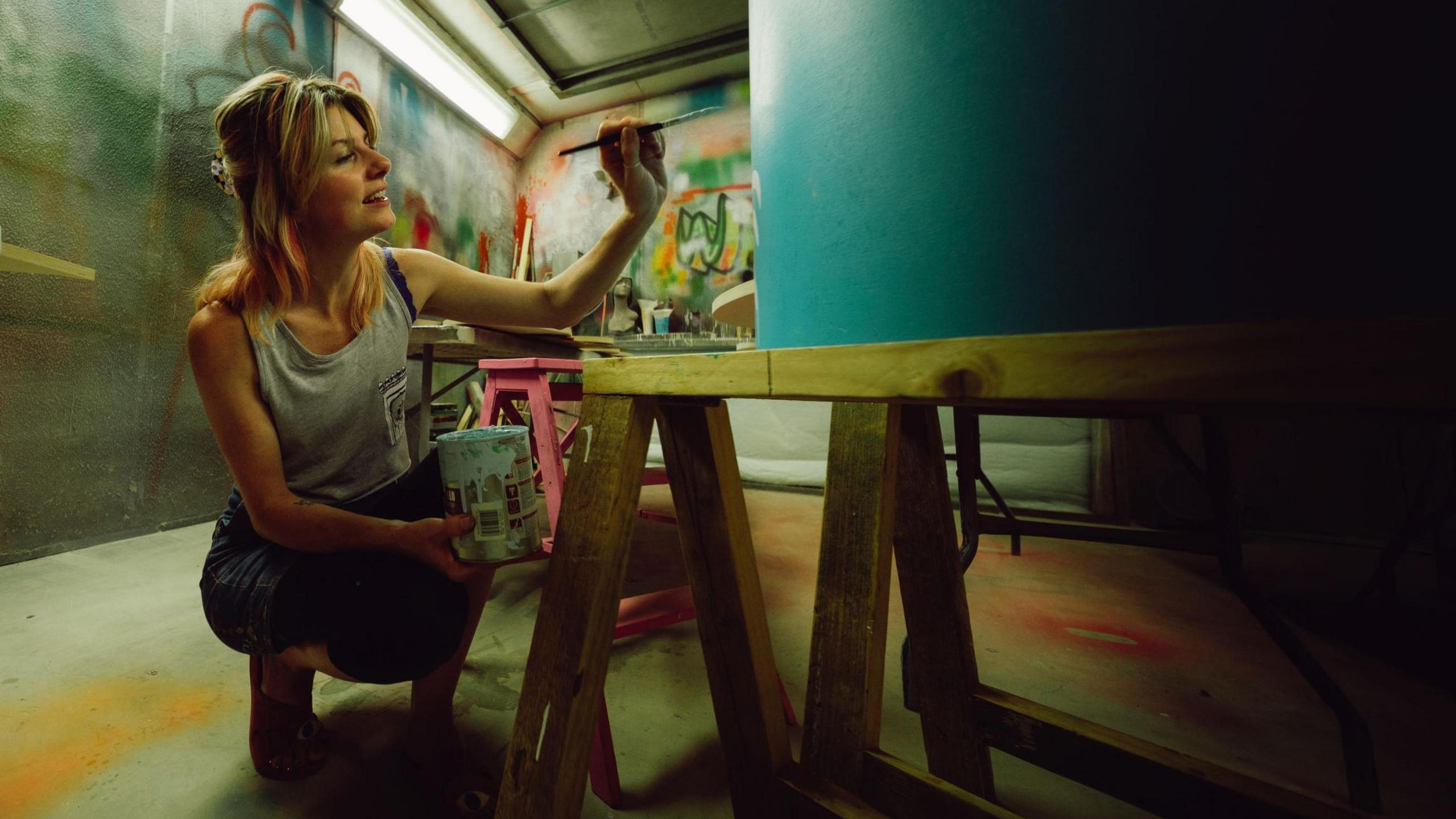 A female artist in jeans and a grey top crouches inside a small warehouse-style room as she paints a design on the side of a cylindrical object.
